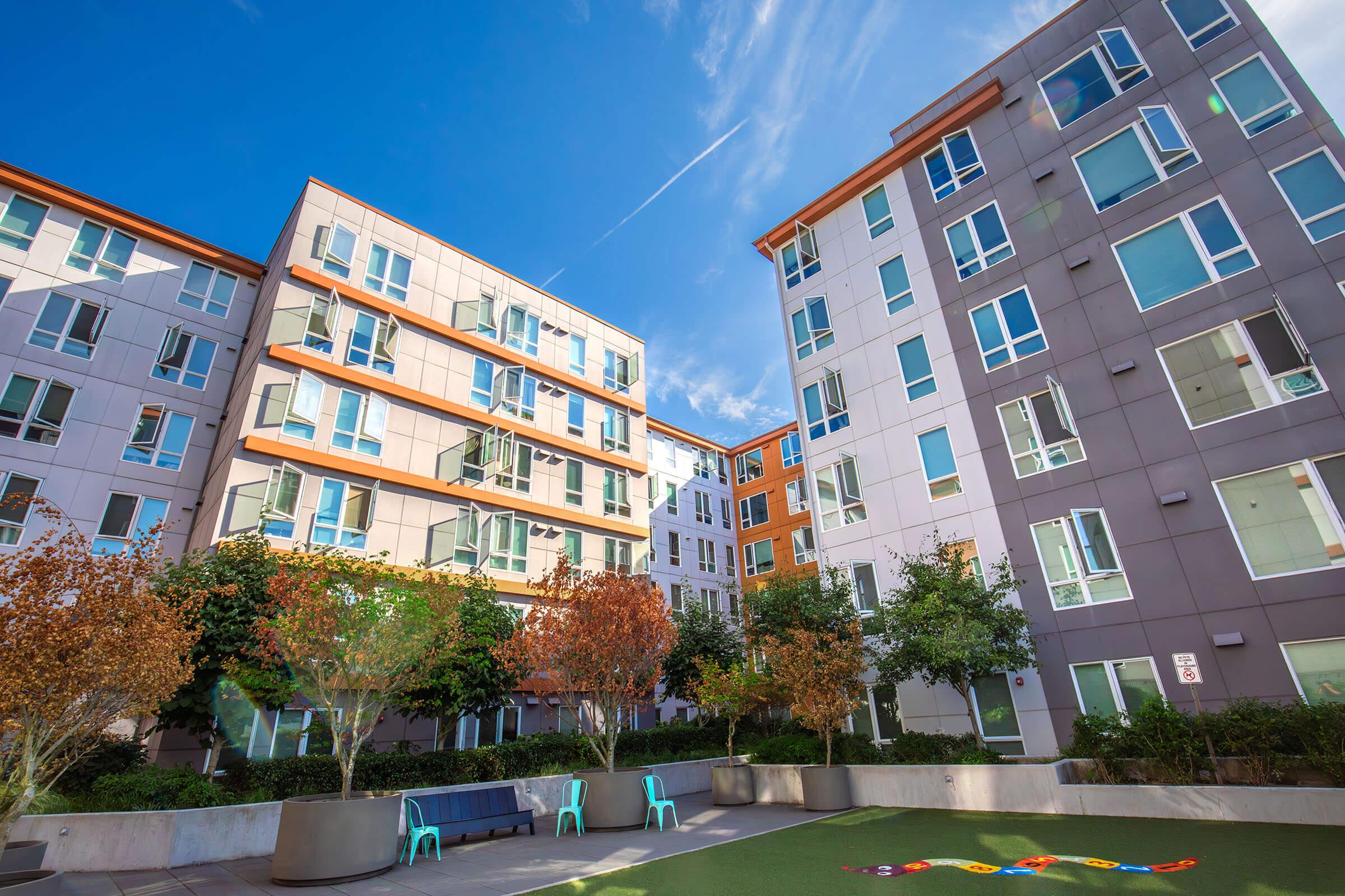 Modern apartment buildings surround a landscaped courtyard with low green grass, colorful chairs, and trees with autumn foliage. The sky is blue with scattered clouds, creating a bright and inviting atmosphere in a residential complex.