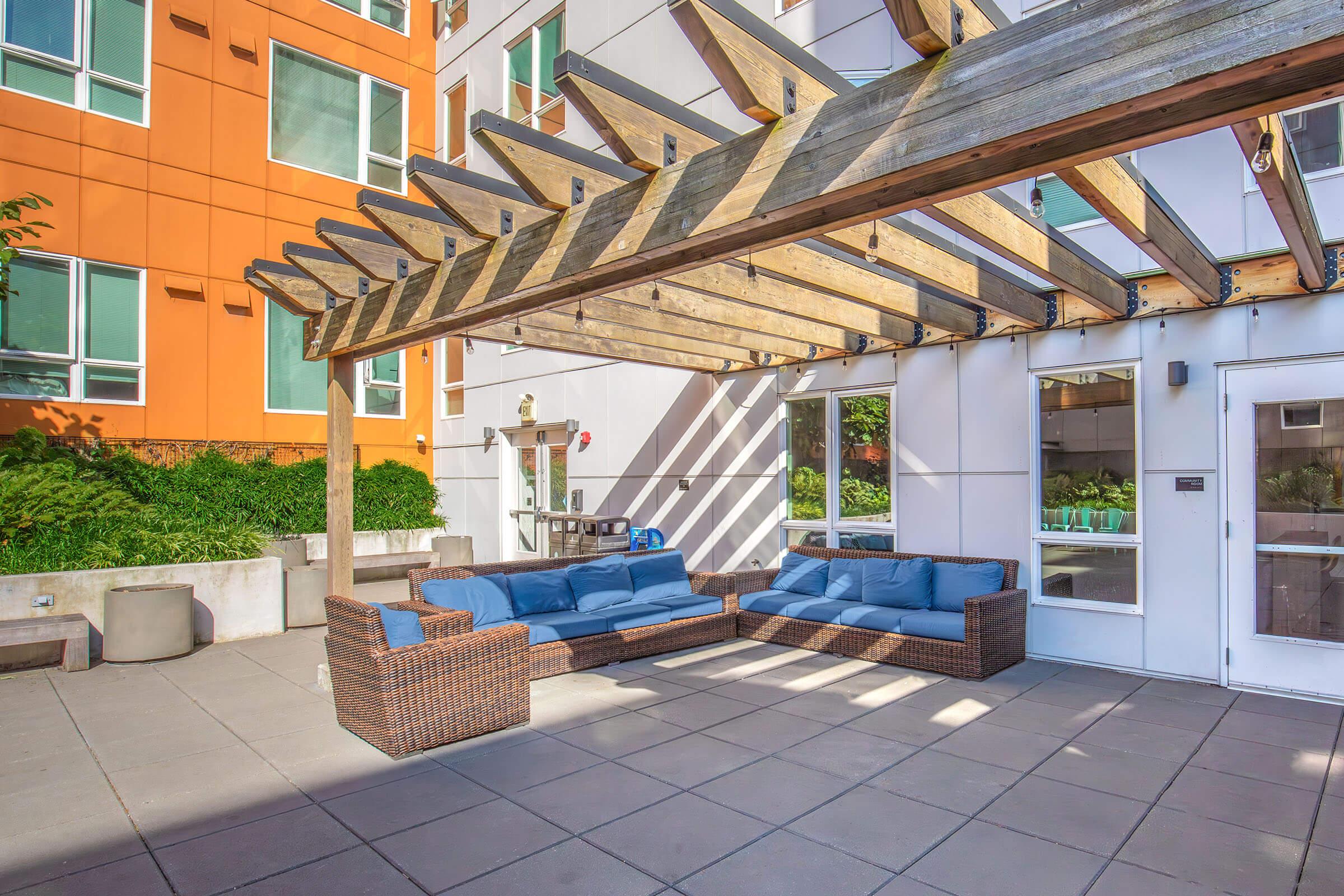 An outdoor seating area featuring a large, brown wicker lounge set with blue cushions, positioned beneath a wooden pergola. The background showcases modern buildings with orange and white facades, and greenery surrounding the space. Sunlight casts shadows across the tiled patio.