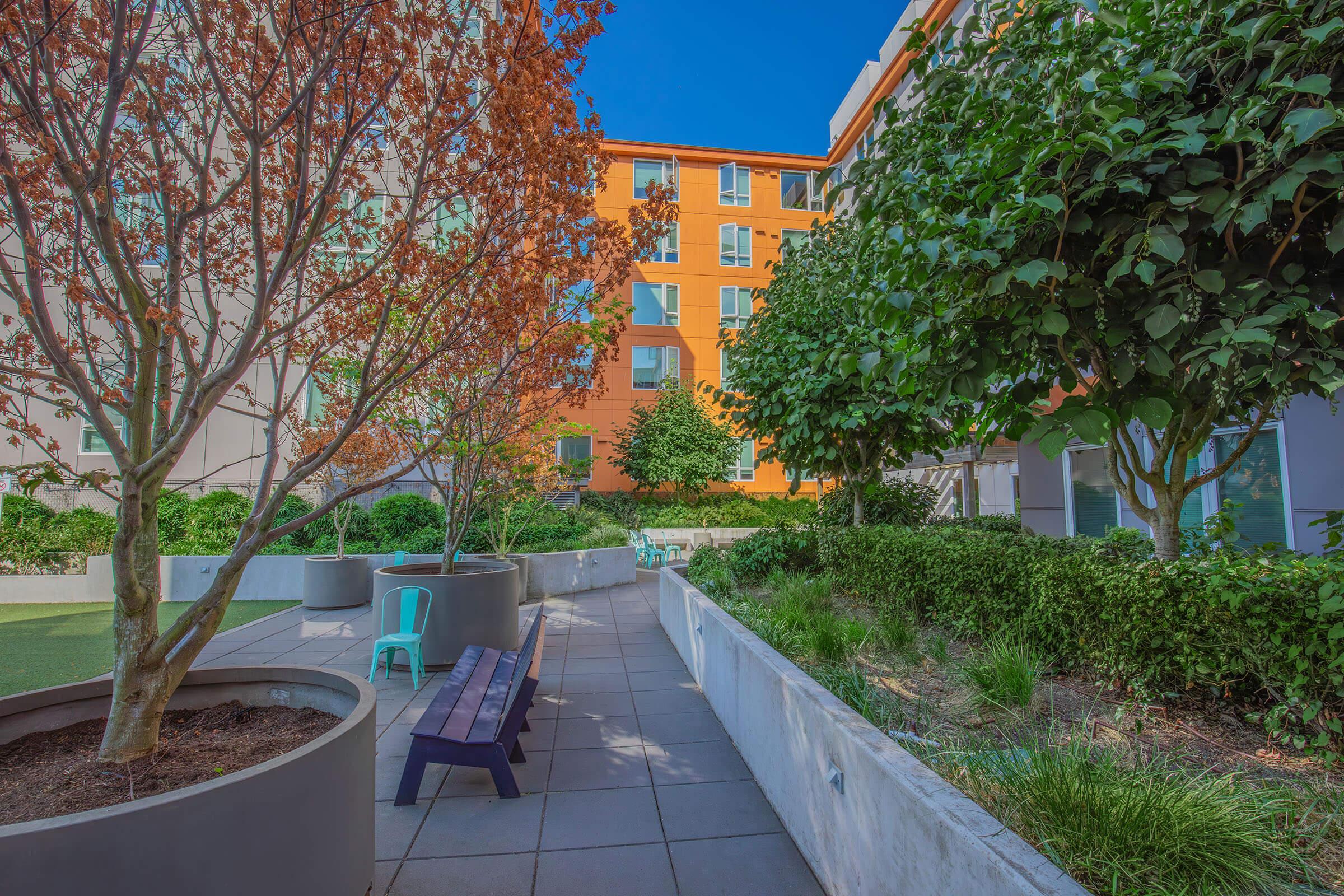 A landscaped courtyard featuring planters with trees and shrubs, a bench, and bright orange building facades in the background under a clear blue sky. The pathway is paved and surrounded by greenery, creating a peaceful outdoor space.