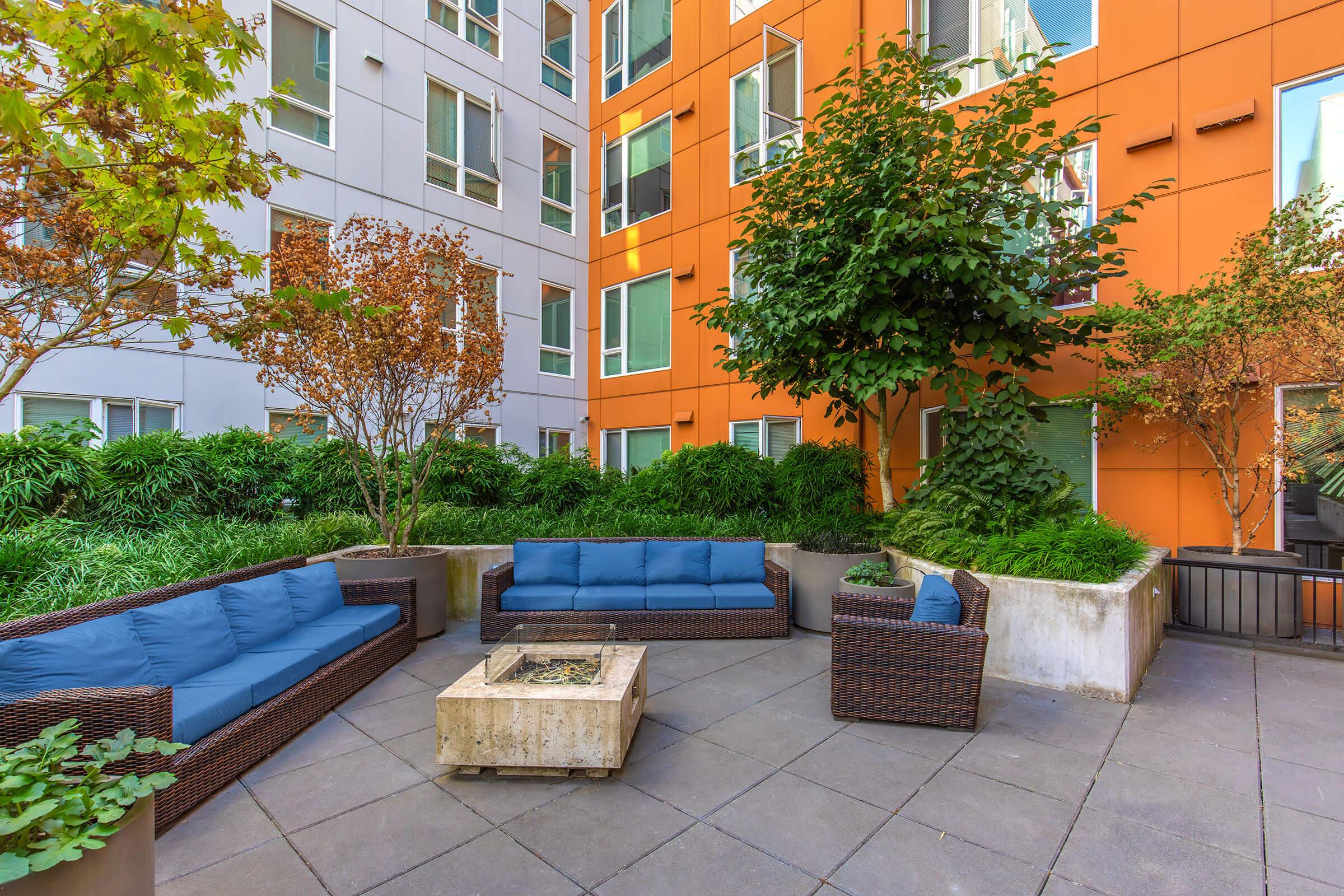 A cozy outdoor lounge area featuring blue cushioned sofas, a central stone fire pit, and surrounding greenery. The backdrop includes a modern building with orange and gray walls, creating a bright and inviting atmosphere. Perfect for relaxation or gathering with friends.