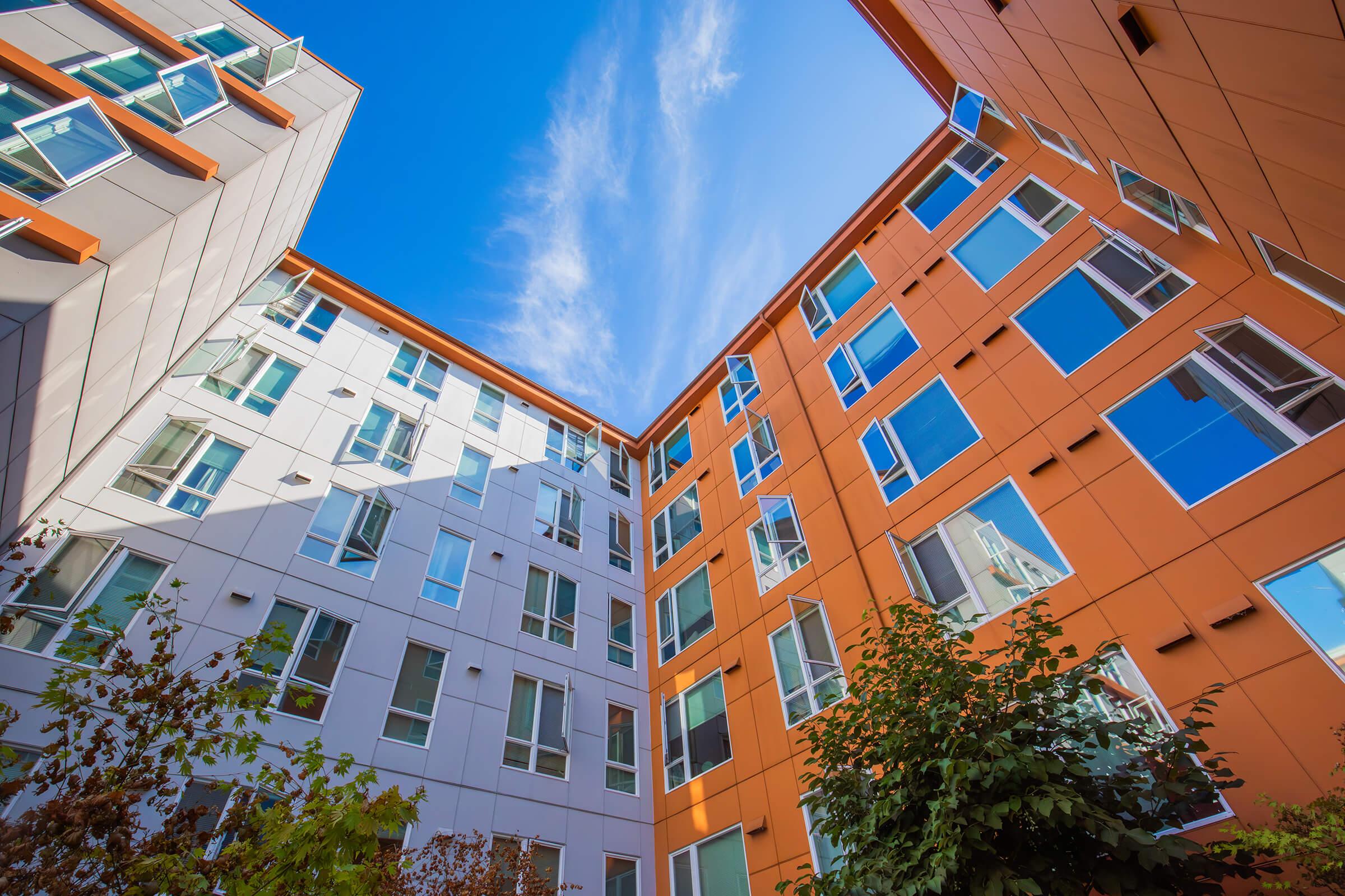 A view looking up at modern apartment buildings with a mix of orange and white facades. The buildings feature numerous windows reflecting the blue sky and scattered clouds above. Lush greenery is visible in the foreground, adding a touch of nature to the urban setting.