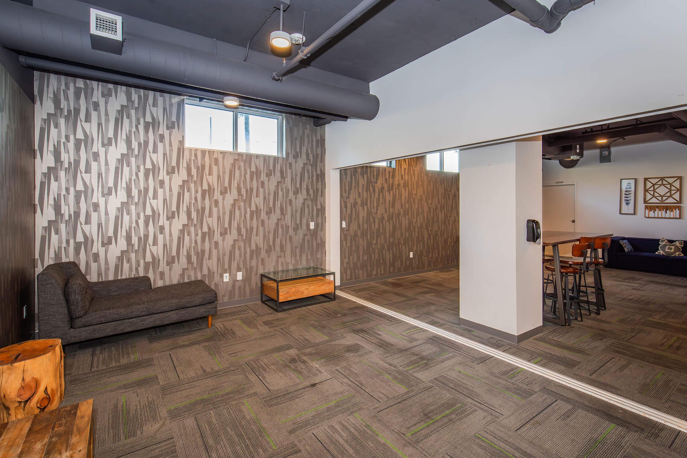 Interior view of a modern room featuring a gray accent wall with a textured pattern, a dark gray sofa, a glass coffee table with a wooden base, and a mirrored wall. The flooring is carpeted with a geometric pattern, and there are large windows providing natural light. The space is minimalistic and contemporary in design.