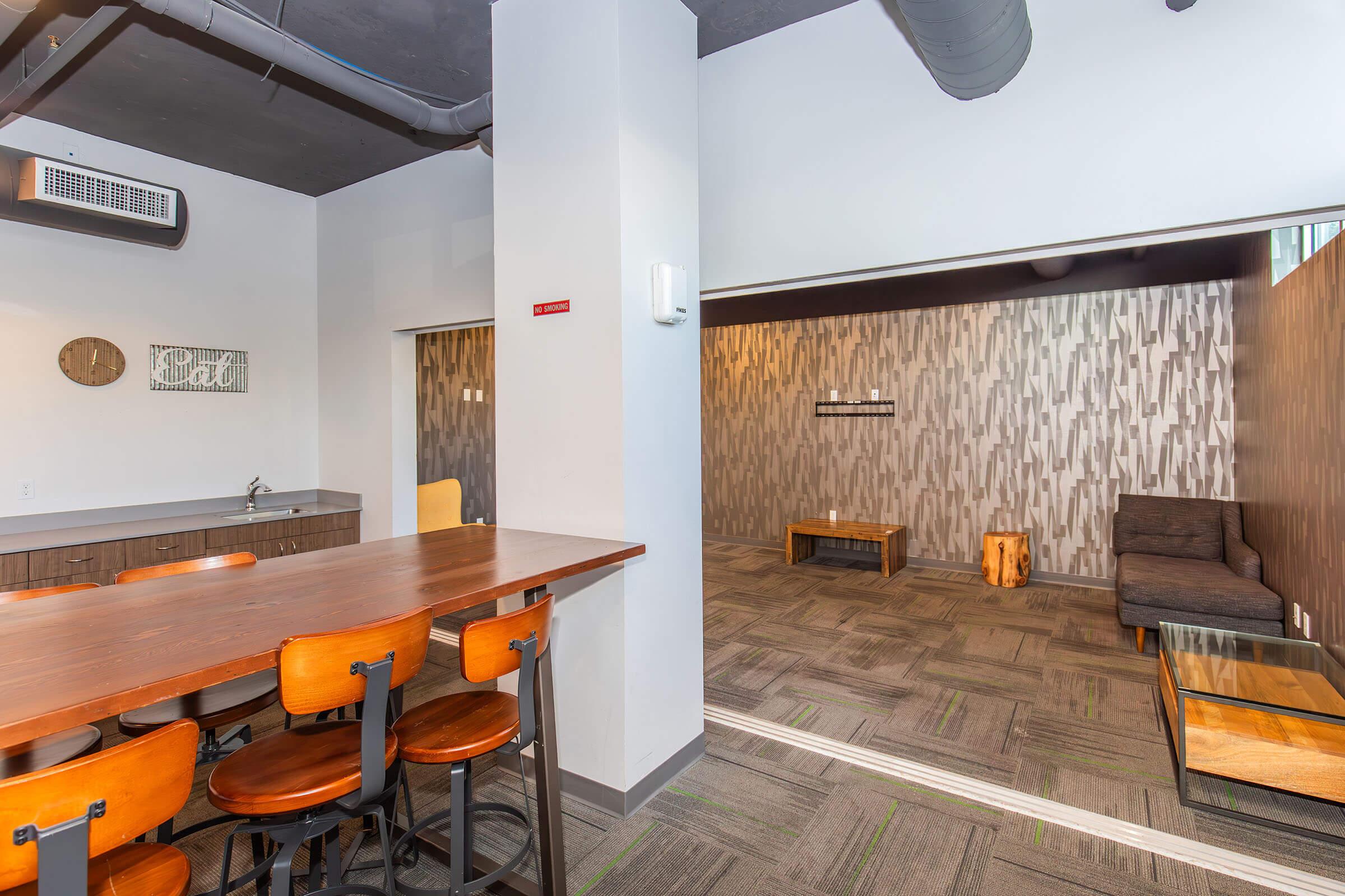 A modern interior featuring a spacious open area with a kitchen counter and bar stools on the left. On the right, there is a cozy seating area with a dark couch and wooden accents, complemented by textured wall paneling. The flooring is carpeted in a neutral tone, enhancing the contemporary design.
