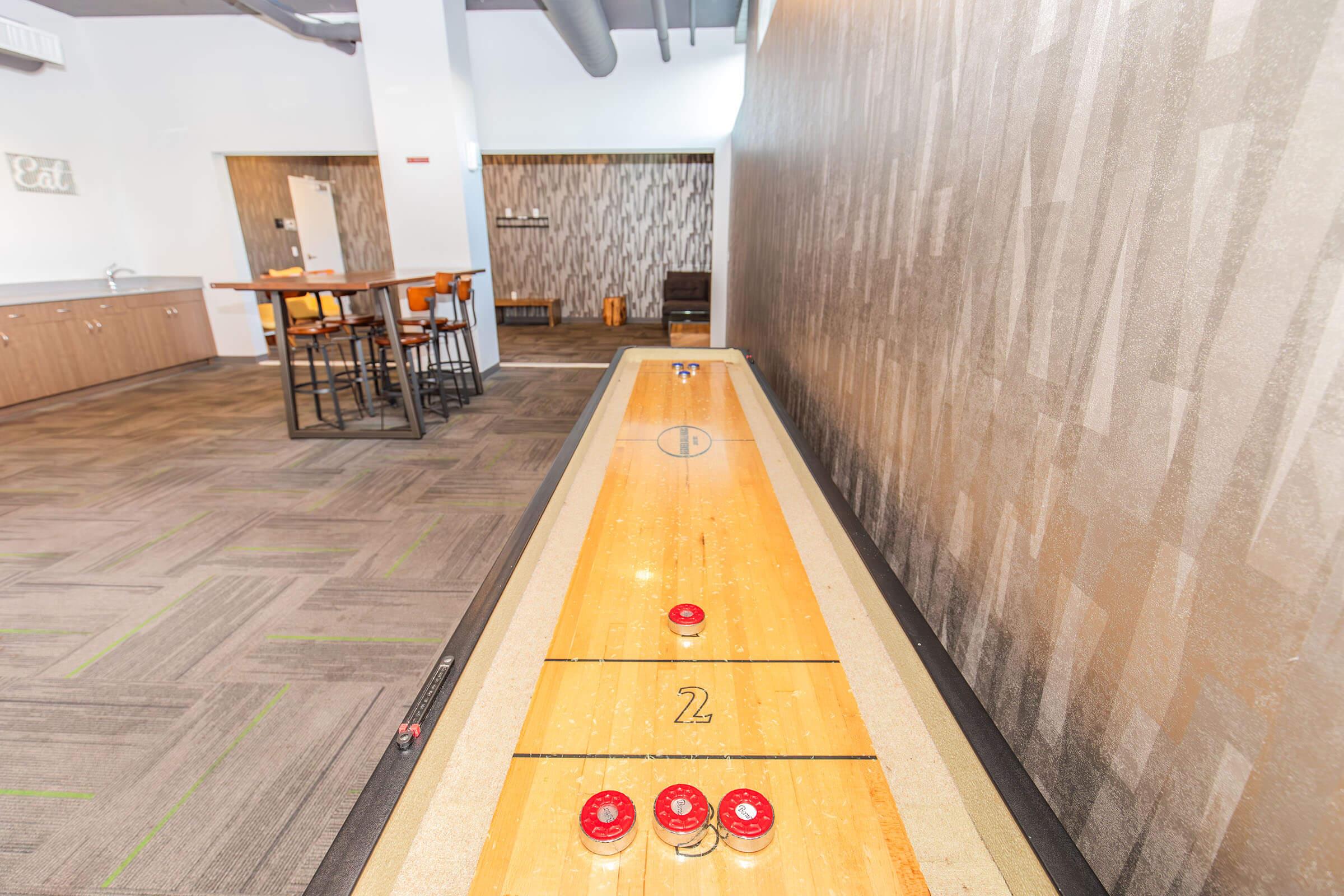 A view of an indoor shuffleboard court with a wooden playing surface, featuring metal discs positioned along the lane. In the background, there are tables and chairs, creating a social environment for players. The walls have a modern texture and color scheme, enhancing the space's inviting atmosphere.