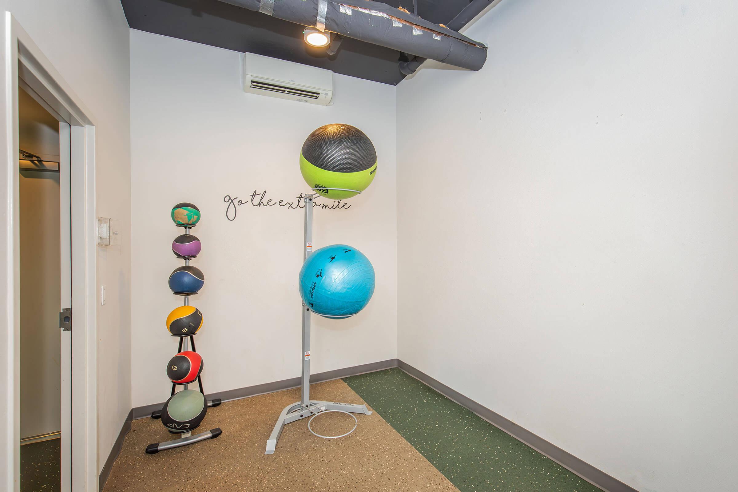 A small fitness room featuring exercise balls of various sizes and colors displayed on a stand. A motivational phrase, "go the extra mile," is written on the wall. The floor is covered with a rubber mat, and there is an air conditioning unit on the ceiling.