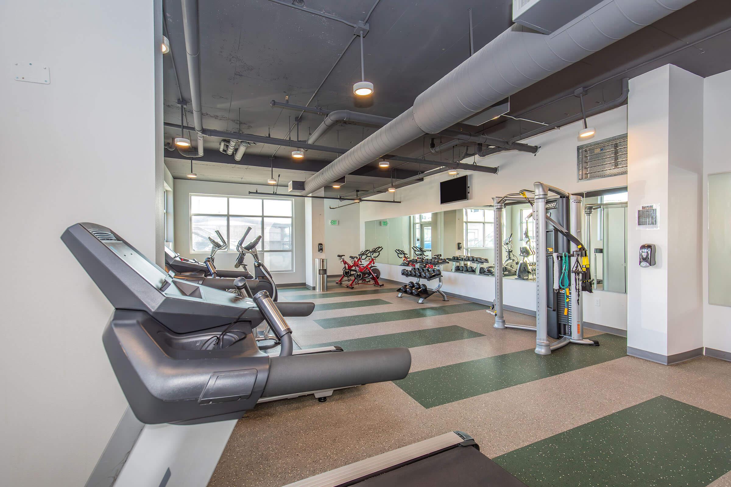 A modern gym space featuring cardio equipment like treadmills and stationary bikes, with weights and resistance machines. Large windows allow for natural light. The flooring has a mix of colors, and mirrors line one wall to enhance the workout environment. Bright overhead lighting completes the setting.