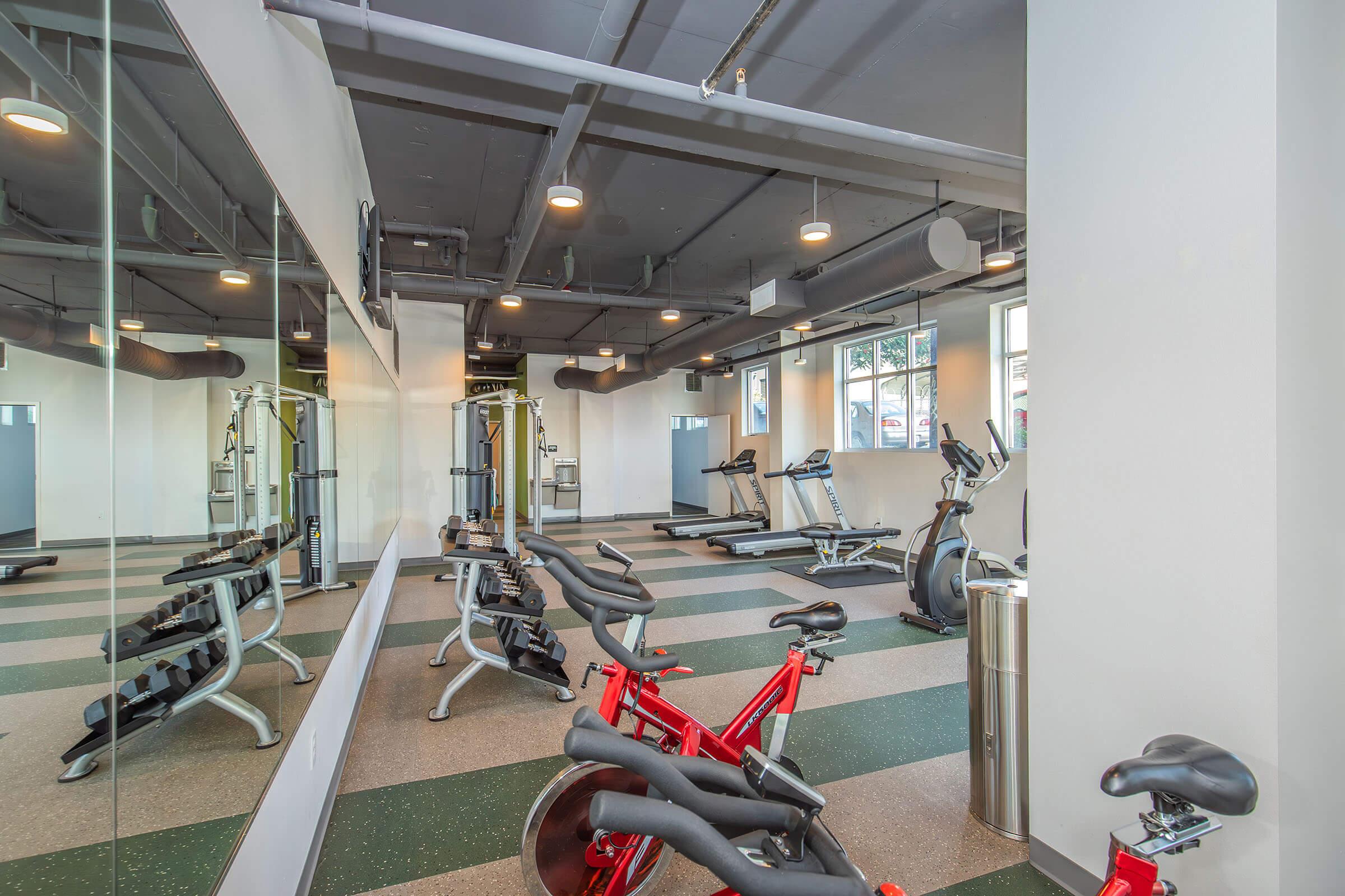 A spacious and modern gym area featuring exercise equipment, including stationary bikes and treadmills. The room has large mirrors on one wall, large windows allowing natural light, and a clean, open layout with green and gray flooring. A small trash bin is visible in the corner.