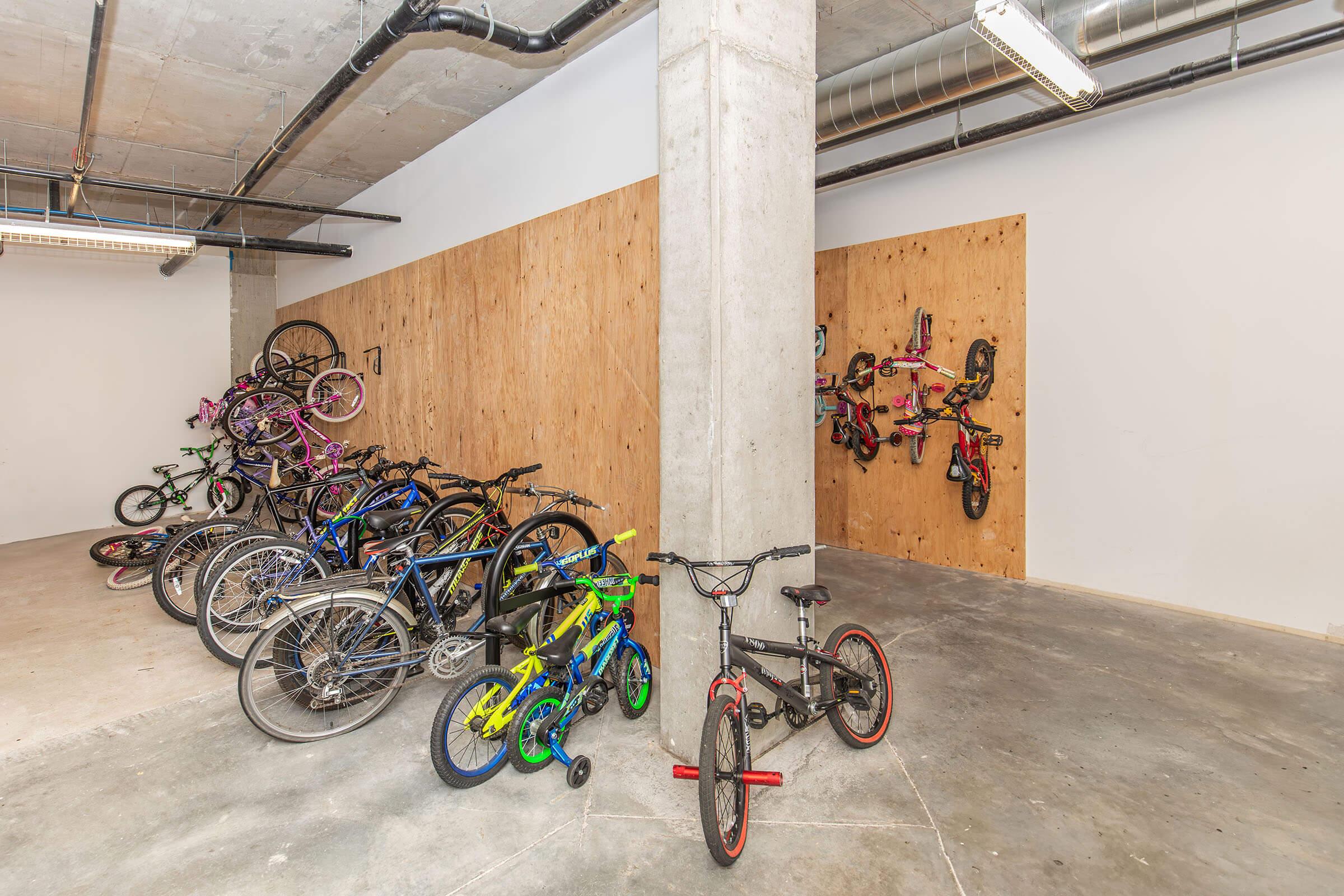 A spacious indoor bike storage area featuring several bicycles of various styles and colors. Bikes are parked on the floor and hung on a wooden wall, showcasing a mix of kids' and adult bikes in a modern, minimalist environment. The concrete walls and flooring create an industrial feel.