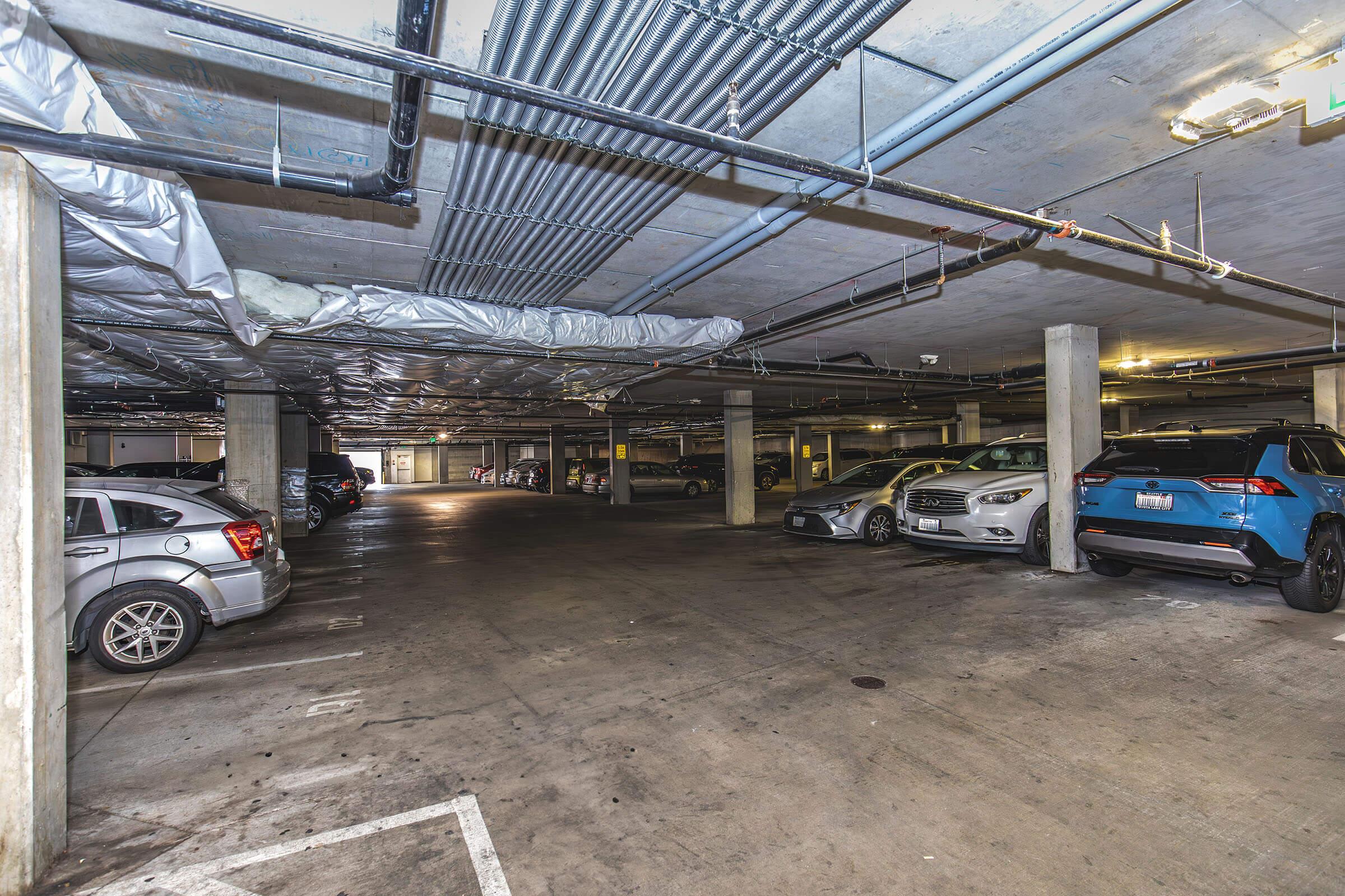 A dimly lit underground parking garage with rows of parked cars. The floor is concrete, and the ceiling has exposed pipes and ductwork. There are several empty parking spaces visible, creating a spacious atmosphere. The scene conveys a typical urban parking environment.