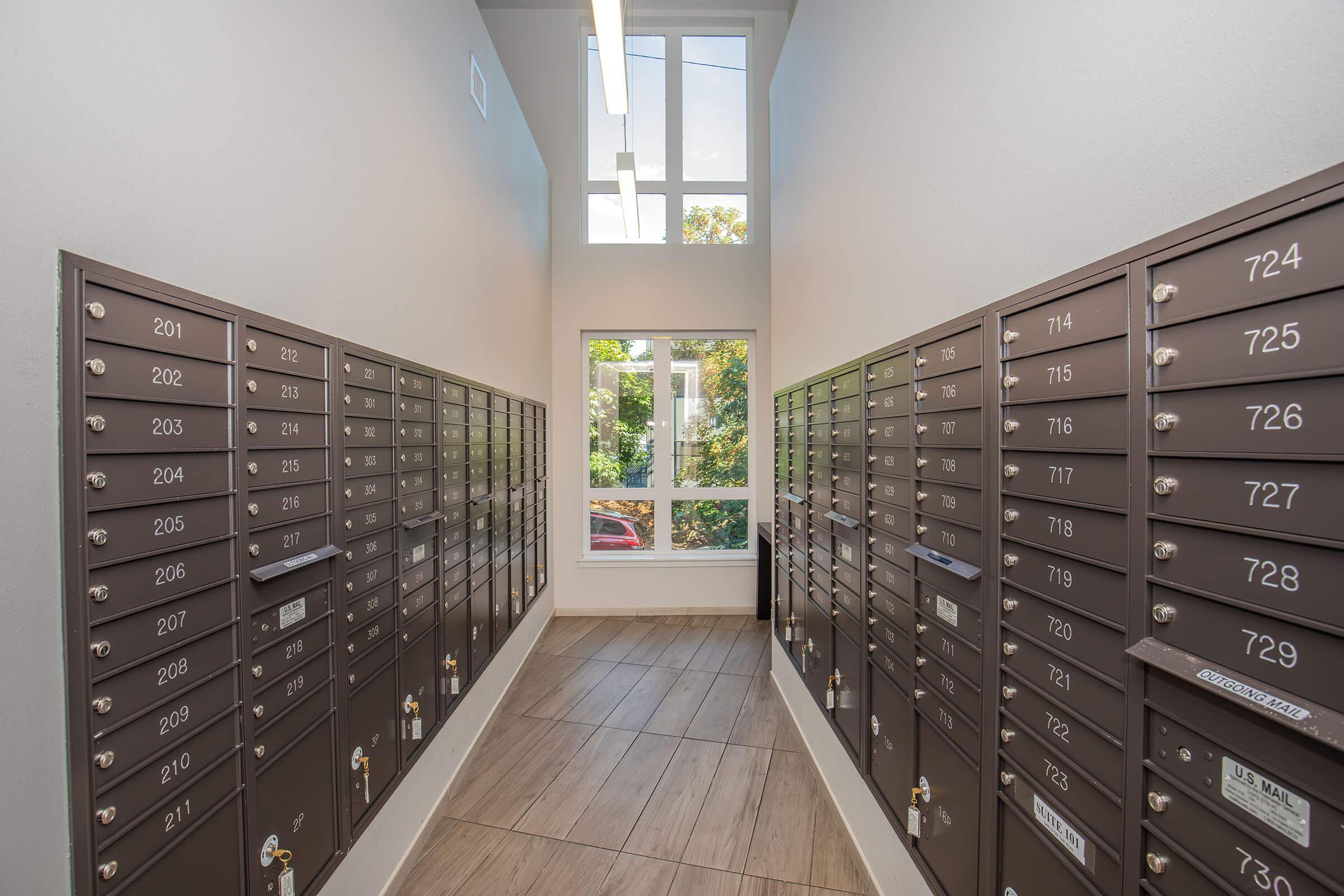 Interior view of a mailroom featuring rows of mailboxes on both sides. The wall-mounted mailboxes are arranged in numerical order, with a modern light fixture and large windows allowing natural light. The floor is tiled, and greenery is visible outside through the windows.