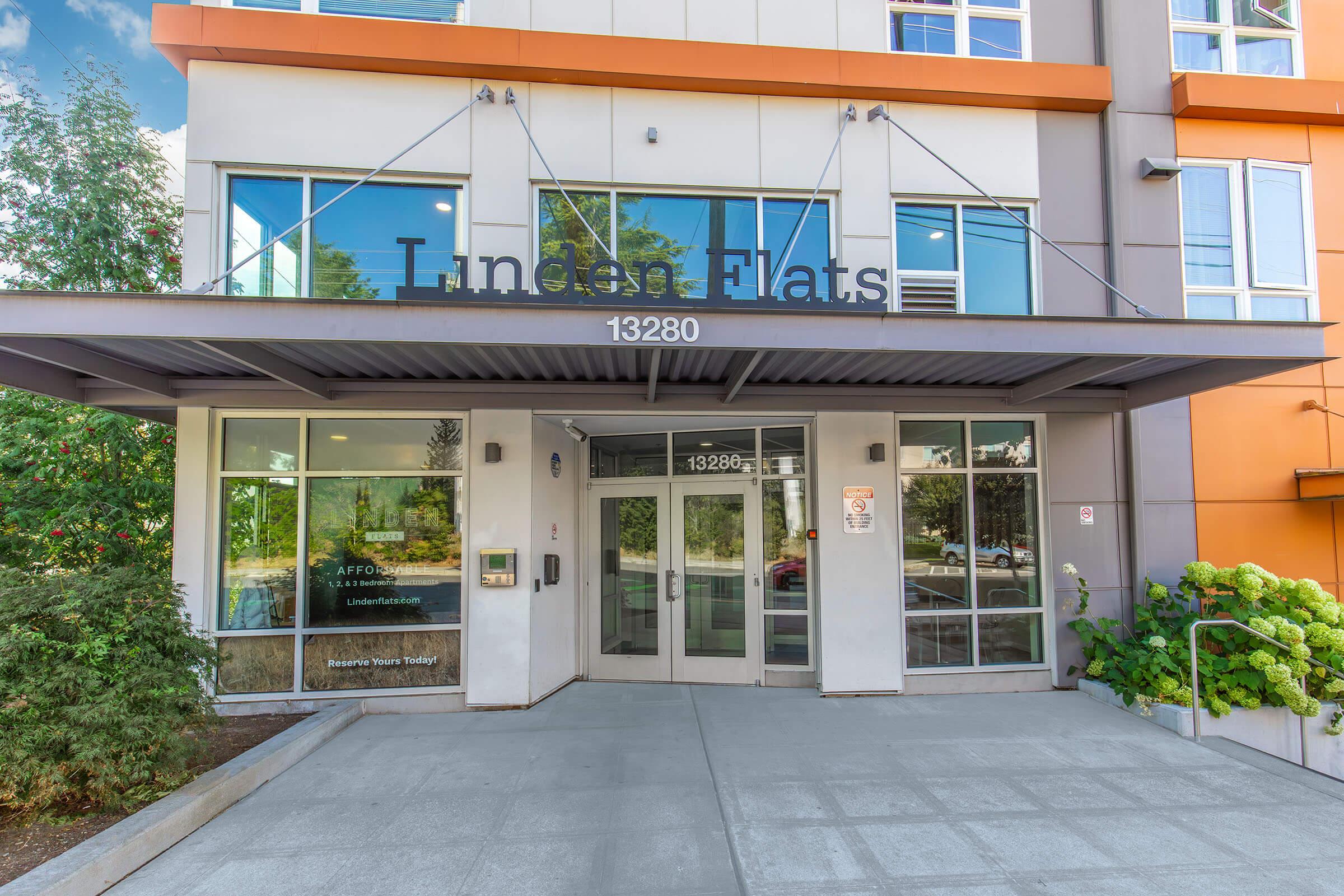 Entrance of Linden Flats at 13280, featuring a modern facade with large glass windows and doors. The surrounding area includes greenery and a concrete pathway, creating an inviting atmosphere. The building has a contemporary design with orange and gray elements.