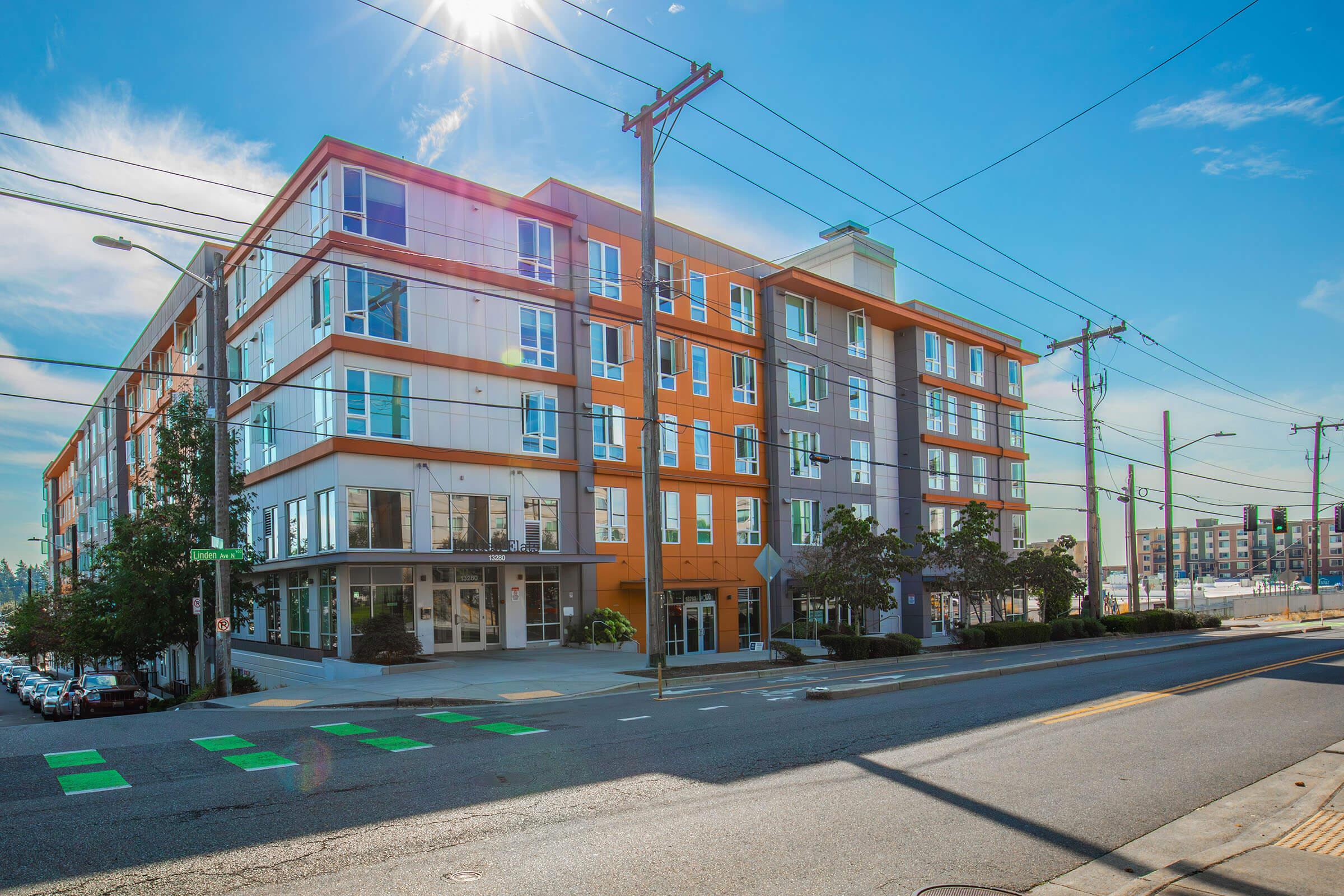 A modern multi-story apartment building featuring a mix of orange, gray, and white exterior panels. The building has large windows, balconies, and is situated on a bustling street with power lines and parked cars in the foreground. The sky is clear with bright sunlight illuminating the scene.