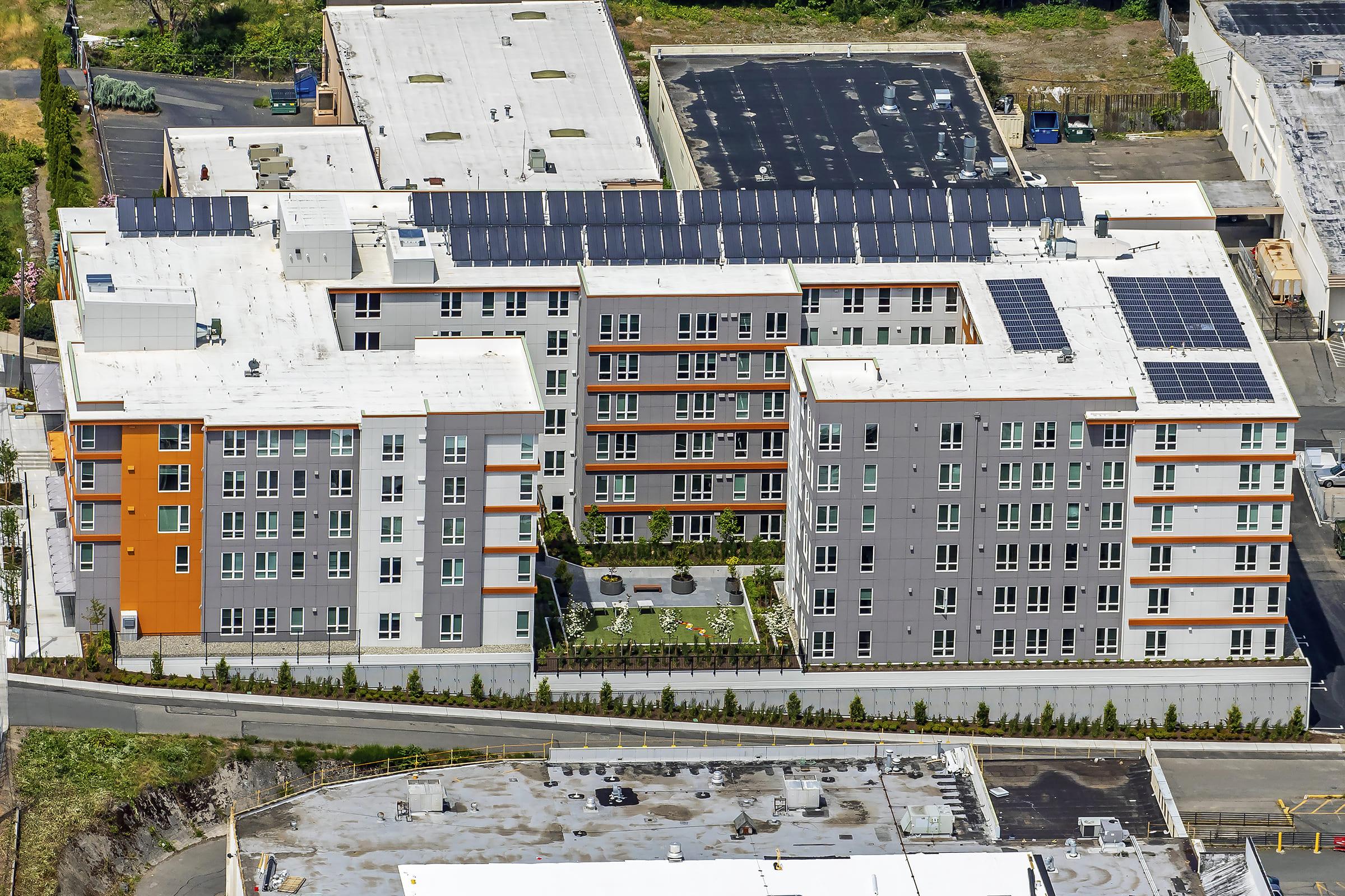 Aerial view of a multi-story residential building featuring modern architecture with a mix of gray and orange facades. The roof is equipped with solar panels, and there is a landscaped area with greenery in the courtyard. Adjacent commercial buildings and parking areas are visible in the background.