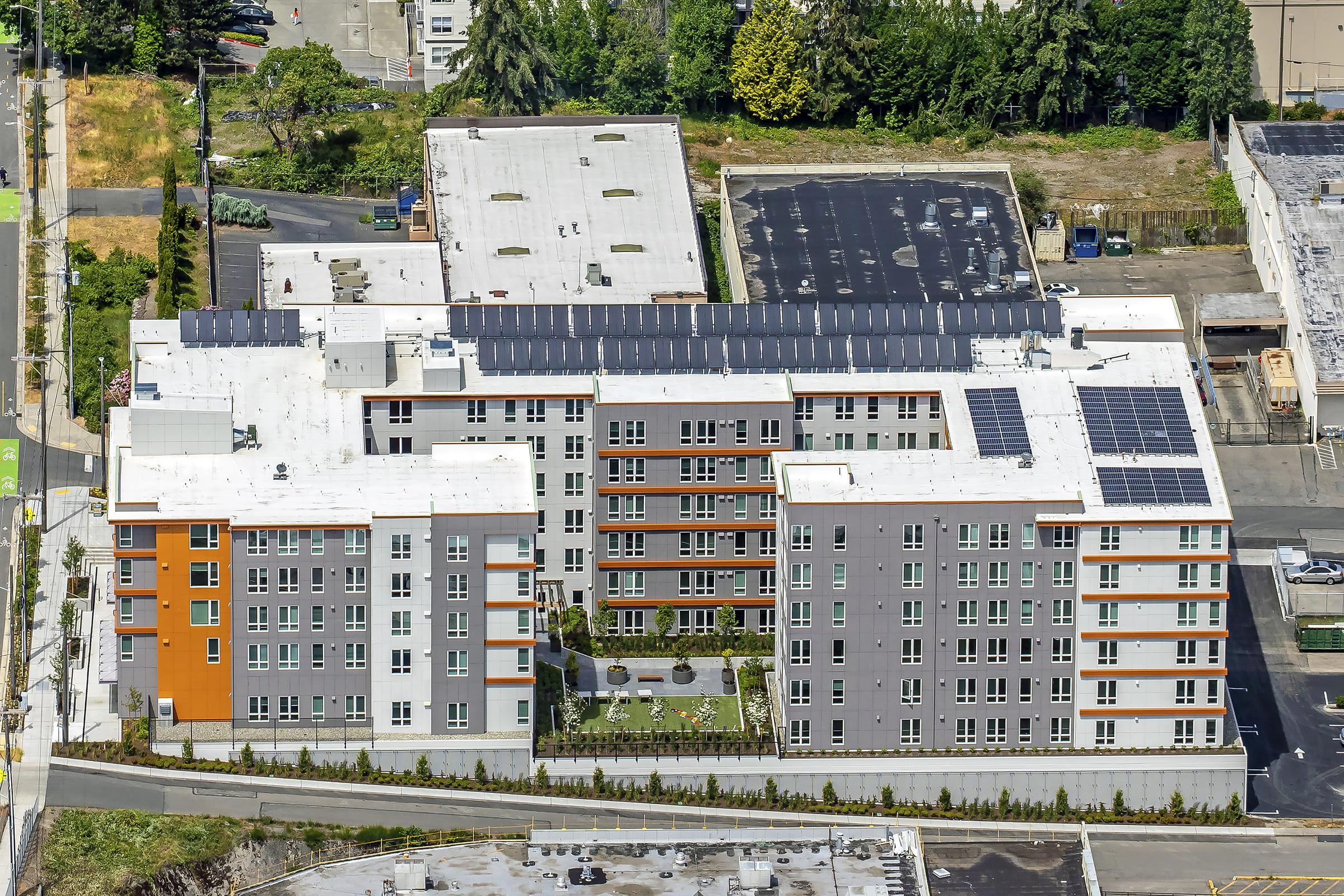 Aerial view of a modern multi-story residential building featuring solar panels on the roof. The building has a mix of colors including gray and orange, with balconies and windows visible. Surrounding areas include green spaces and a paved surface.