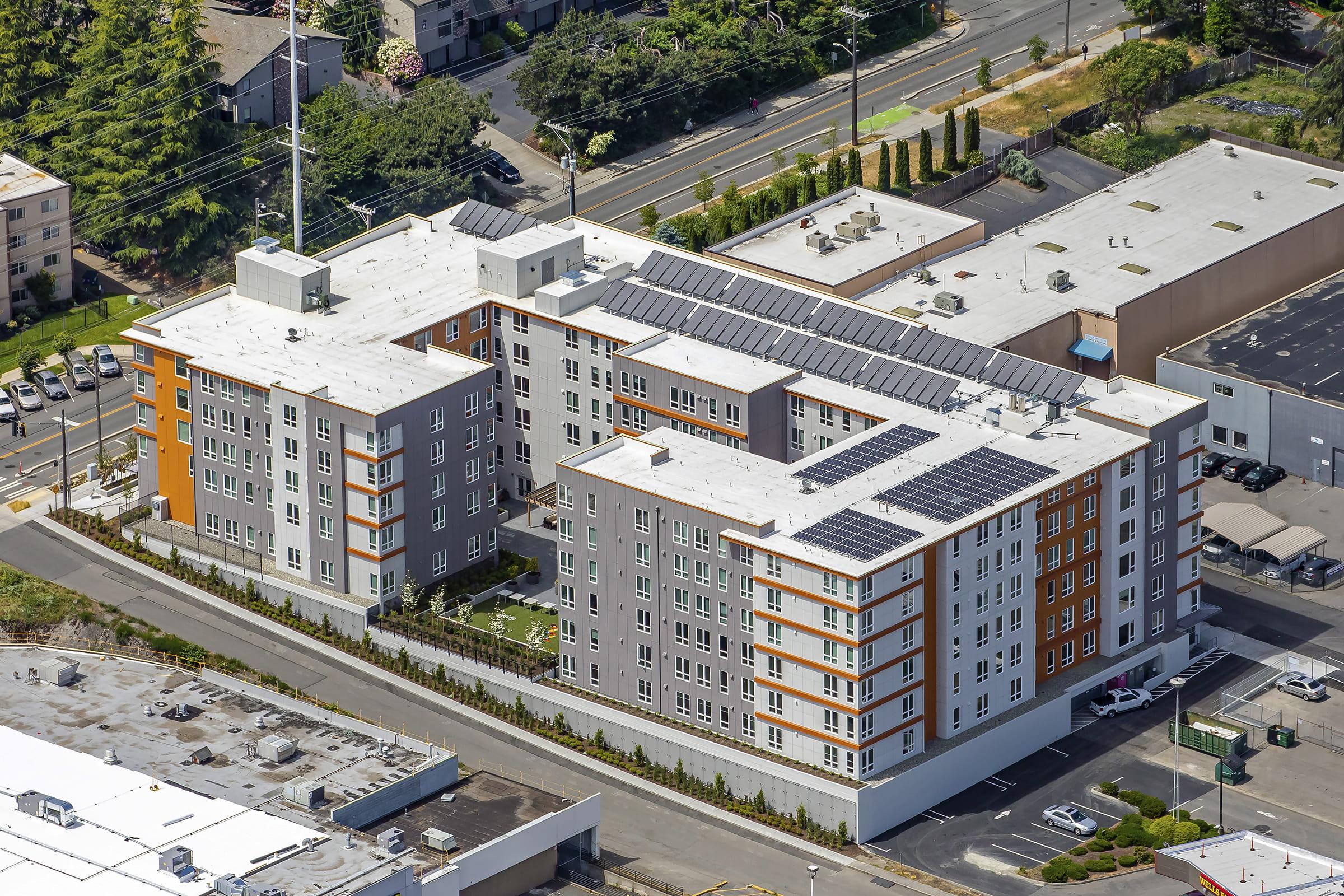 Aerial view of a modern apartment community featuring multiple stories, a mix of gray and orange exterior colors, solar panels on the roof, and landscaped areas. Surrounding the building are parking lots and nearby trees, with neighboring structures and roads visible in the background.