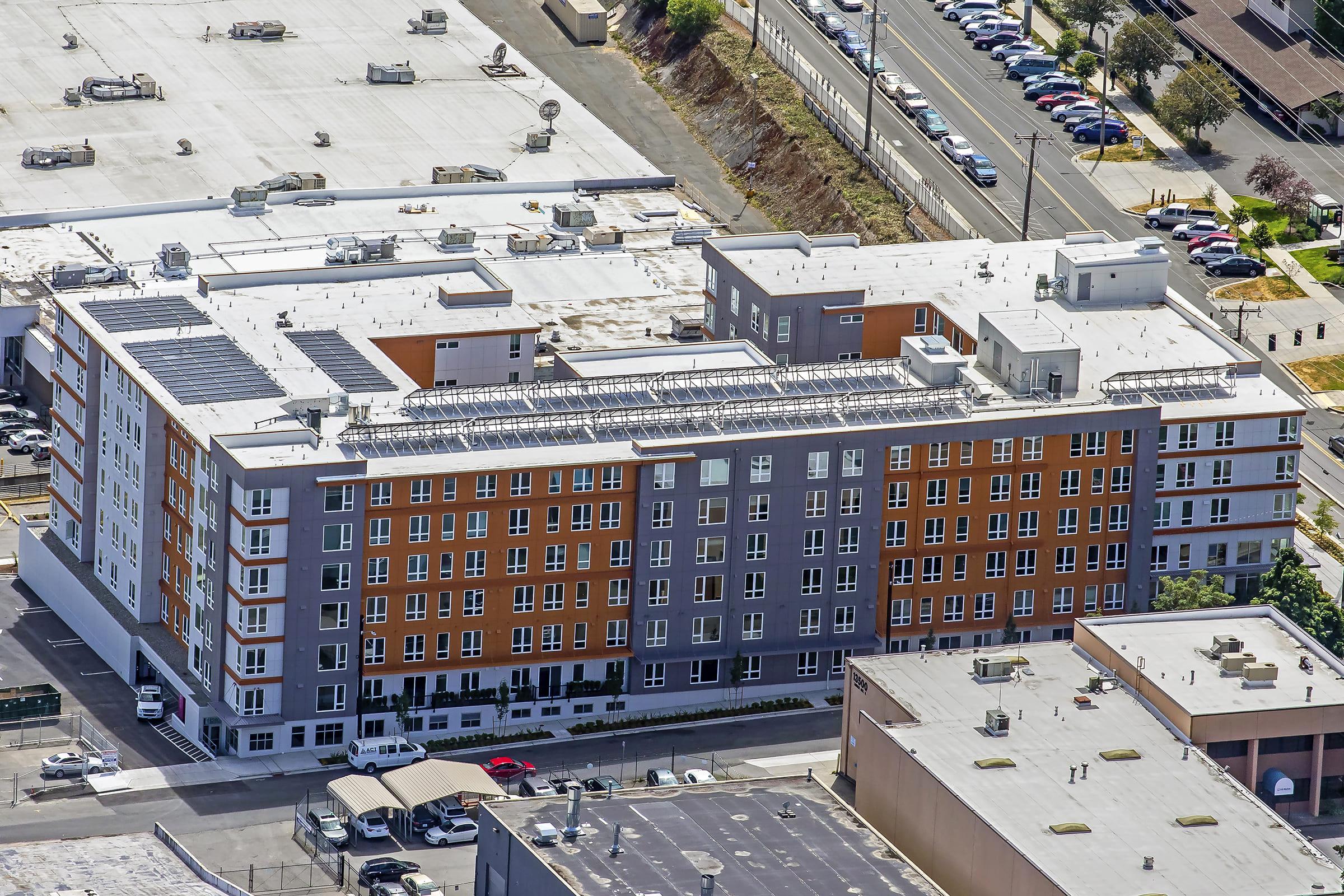 Aerial view of a multistory residential building with a modern design featuring orange and gray facades. The rooftop has solar panels, and the surrounding area includes parking spaces and other buildings. The scene shows a combination of urban architecture and infrastructure.