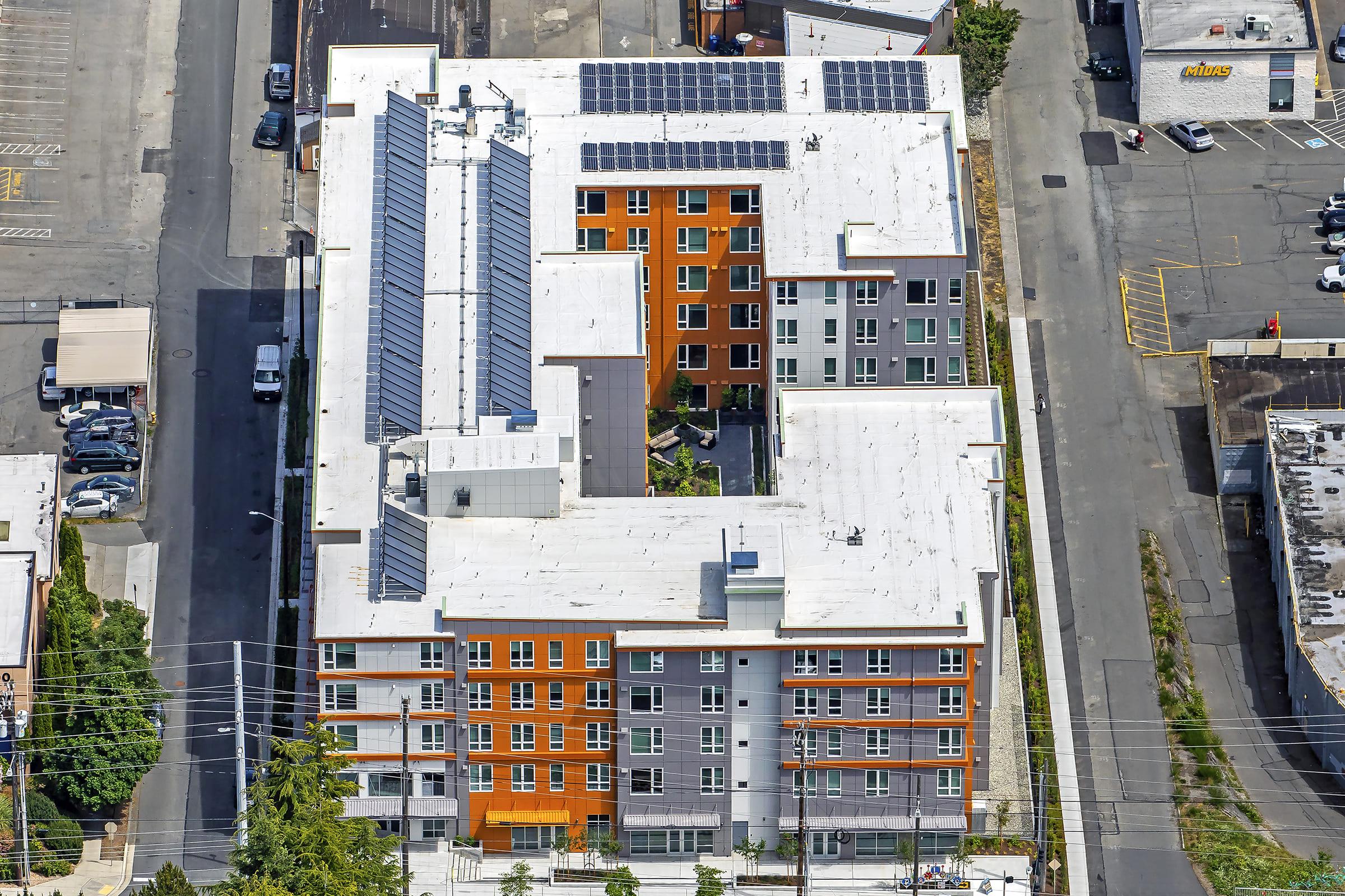 Aerial view of a modern multi-story residential building featuring solar panels on the roof. The building has a combination of orange, gray, and white exterior finishes, with a central courtyard visible. Surrounding the building are roads and parking areas, indicating an urban setting.