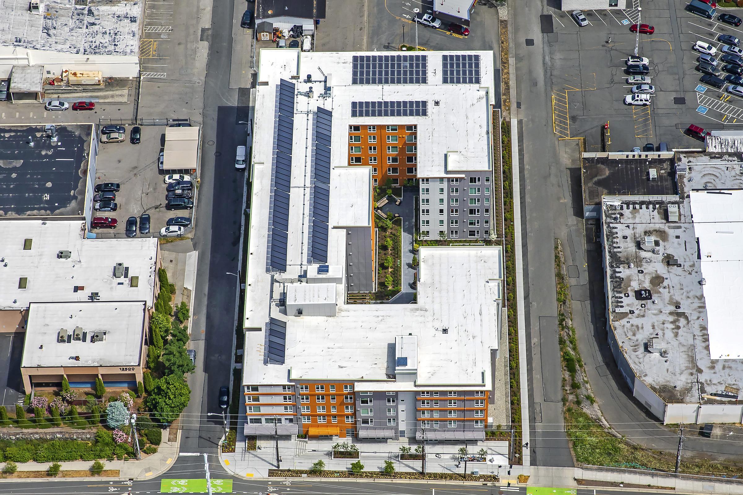 Aerial view of a modern multi-story building complex featuring a flat roof with solar panels. The structure includes colorful facades and an interior courtyard with landscaping. Surrounding areas show parking lots and nearby commercial buildings, along with streets bordering the property.