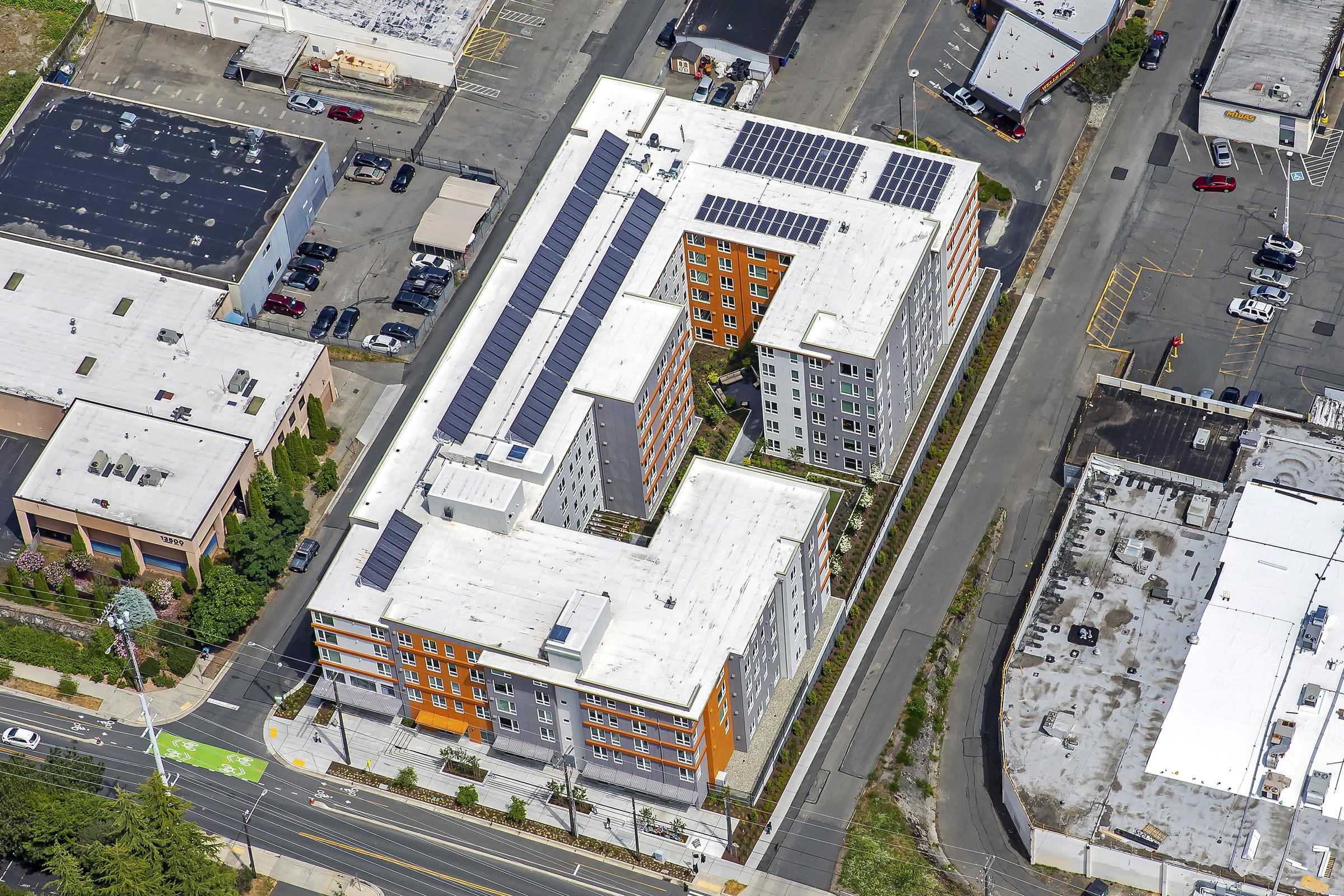 Aerial view of a modern multi-story residential building featuring flat roofs with solar panels. Surrounding structures include commercial buildings and parking areas, with tree-lined streets nearby. The building showcases a combination of orange and grey exterior colors, surrounded by various vehicles parked along the road.