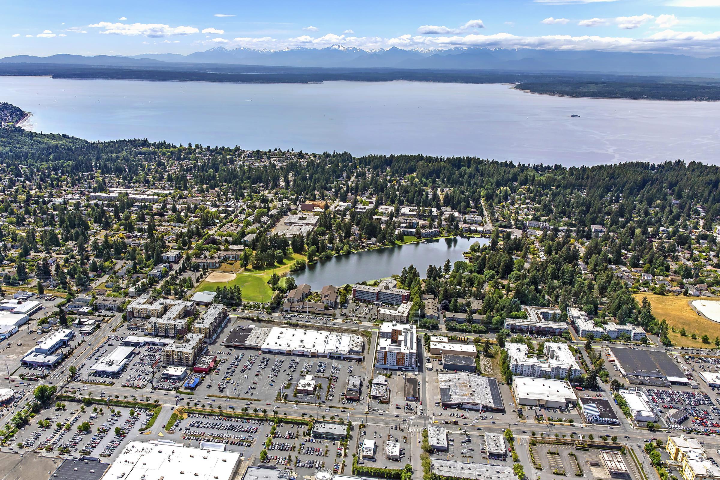 Aerial view of a coastal town overlooking a large body of water with mountains in the background. The image reveals residential areas, commercial buildings, and a green park by a small lake amidst a blend of urban and natural scenery. Clear blue skies enhance the vibrant landscape.