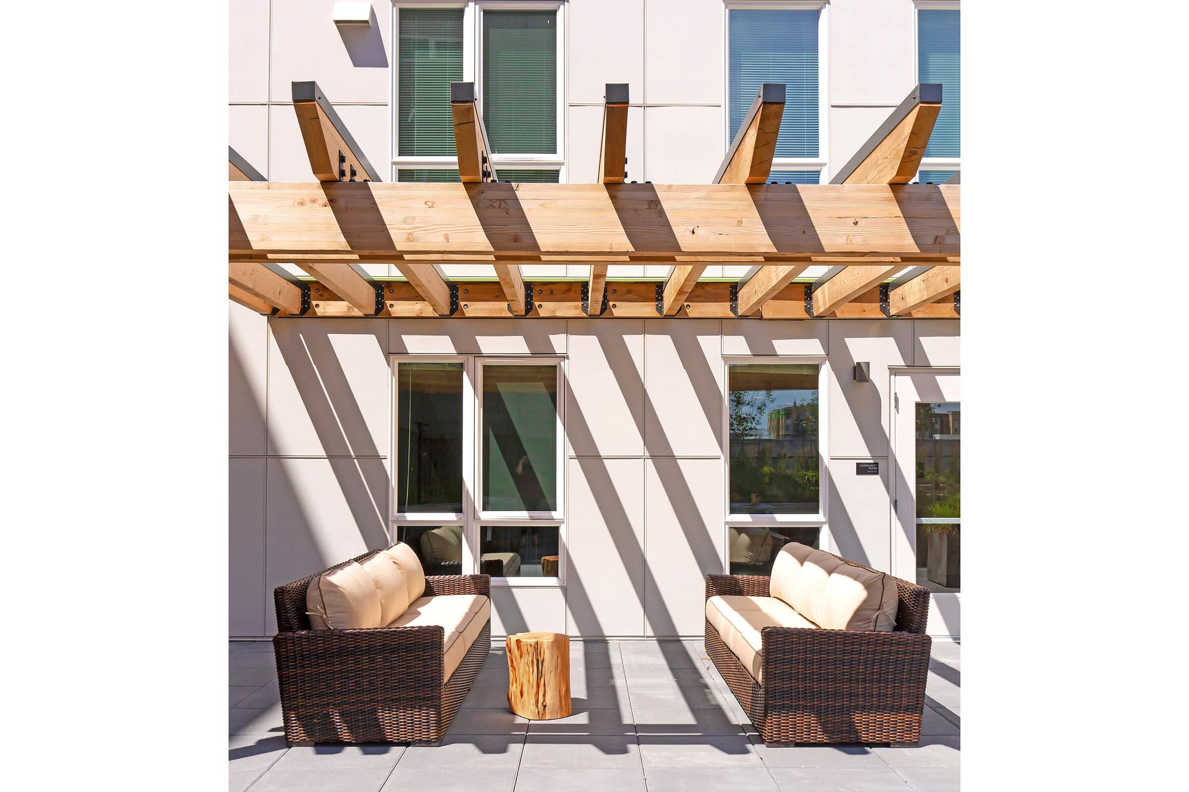 Outdoor seating area featuring two wicker sofas with cushioned seats, positioned under a wooden pergola. A small wooden side table sits between the sofas, and sunlight casts shadows on the wall behind them. The setting has a modern, relaxing feel, ideal for casual gatherings.