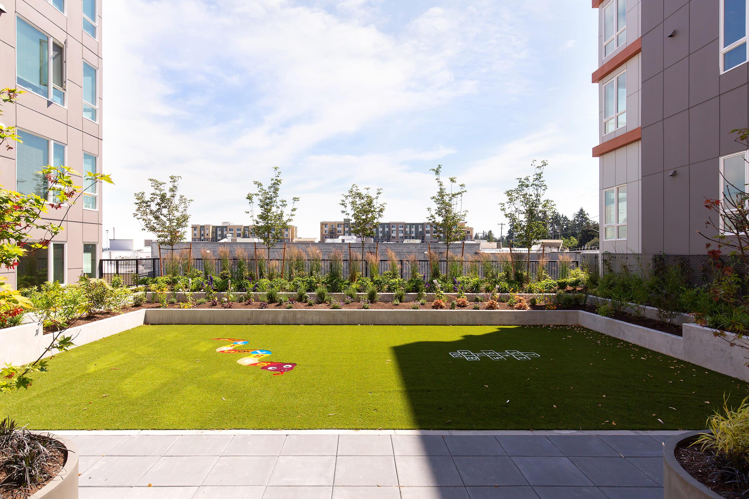 A vibrant outdoor courtyard featuring a green artificial turf area with colorful hopscotch markings. Surrounding the turf are neatly arranged flower beds and small trees, with modern buildings visible in the background under a clear blue sky.