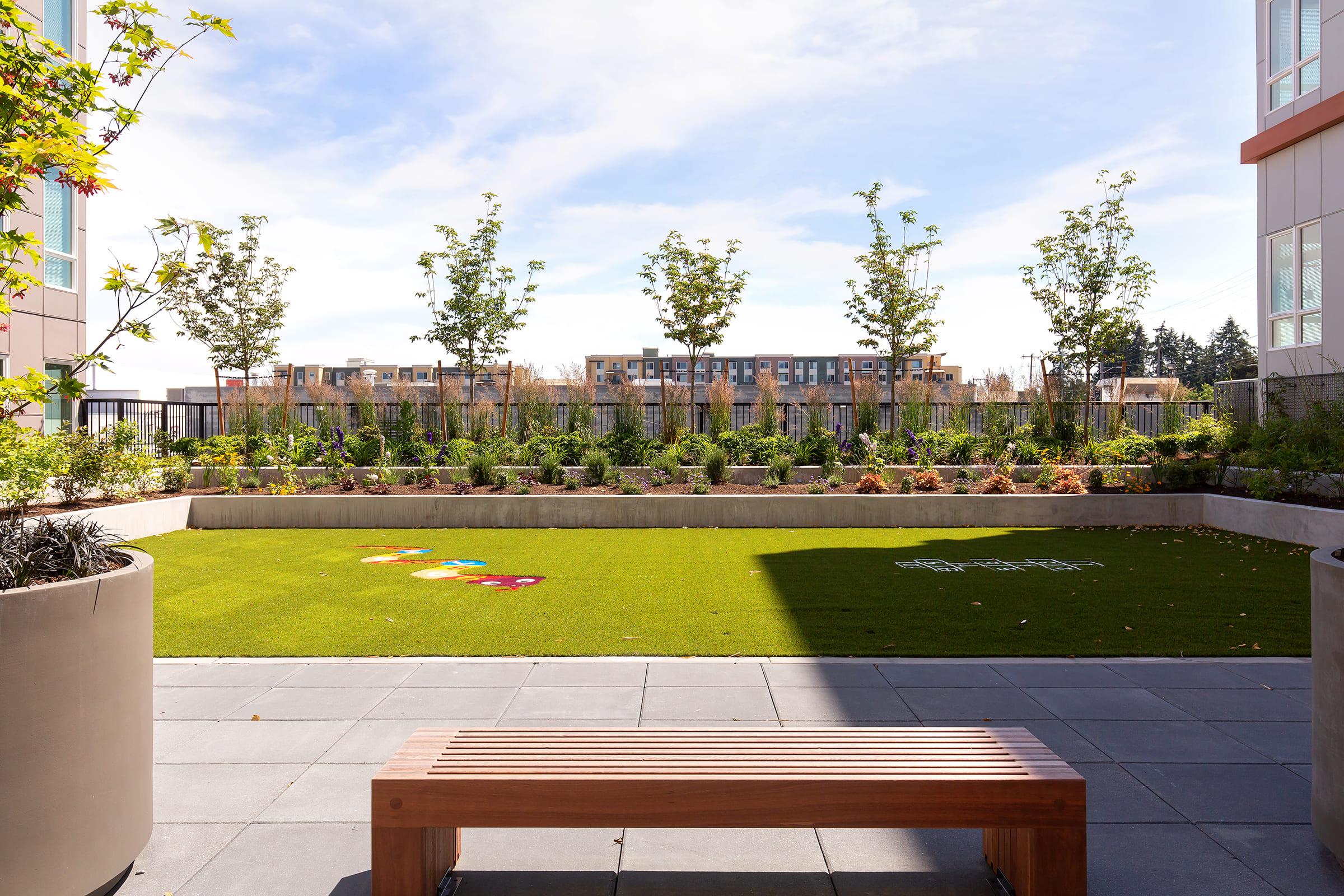 A landscaped outdoor area featuring green grass, neatly arranged flower beds, and young trees under a clear blue sky. A stone path leads through the space, and a wooden bench is placed in the foreground, providing a comfortable spot to enjoy the garden view.