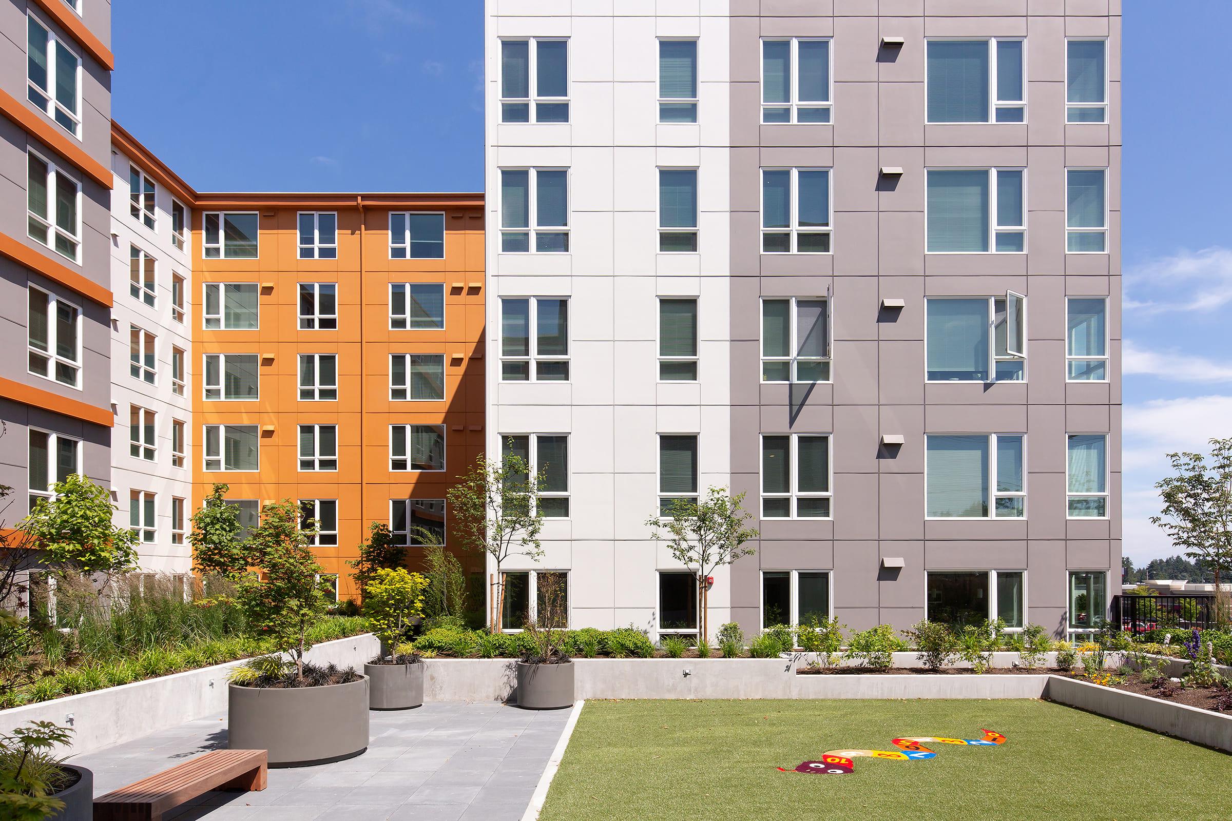 Modern multi-story buildings in varied colors surround a landscaped courtyard. The courtyard features green grass, planters with small trees, and colorful children's toys scattered on the ground. Bright blue sky and sunlight enhance the inviting atmosphere of the area.