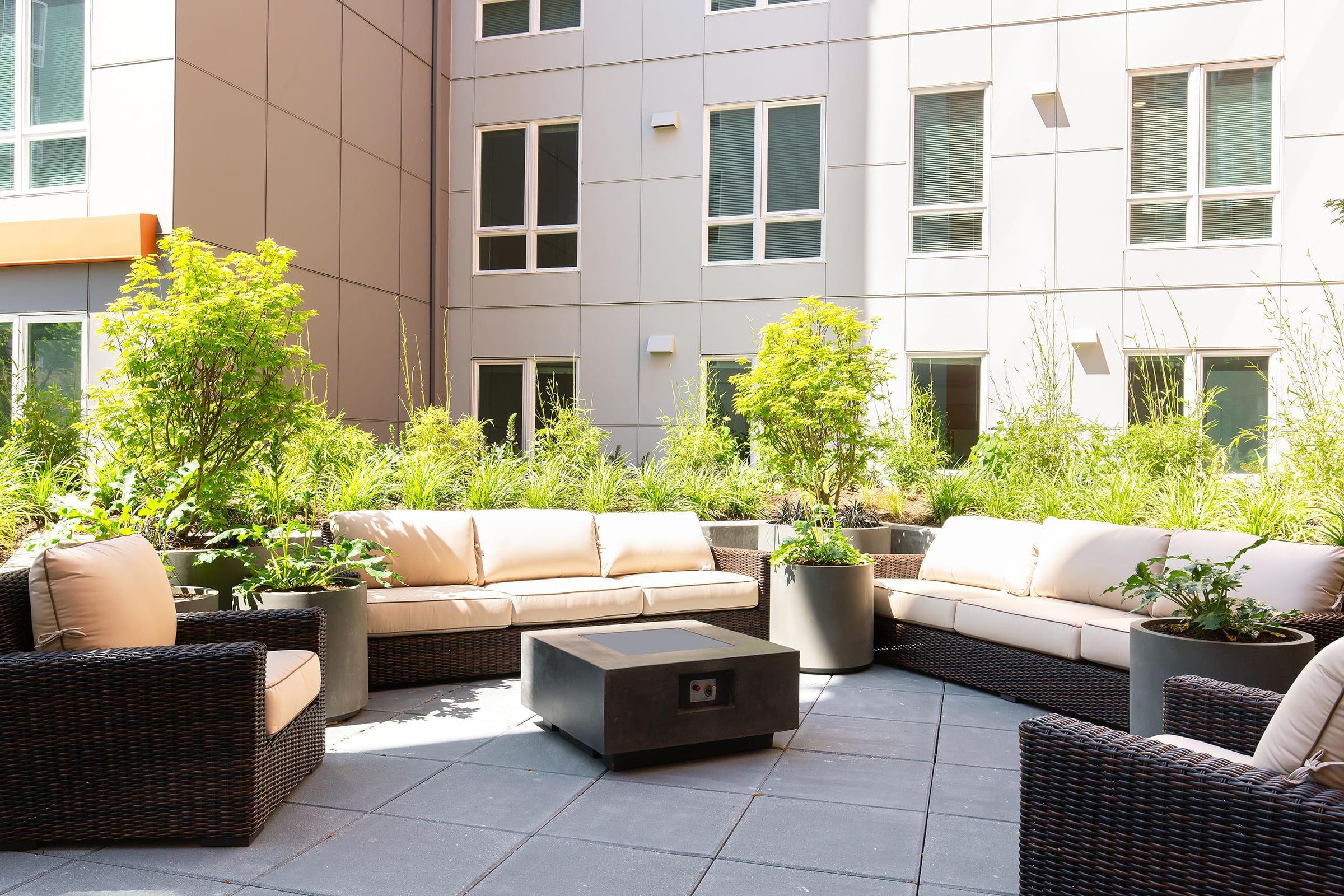 A stylish outdoor seating area featuring beige upholstered sofas and armchairs with dark wicker frames. A modern coffee table sits in the center, surrounded by lush greenery in planters. The backdrop consists of contemporary building windows, creating a serene and inviting atmosphere.