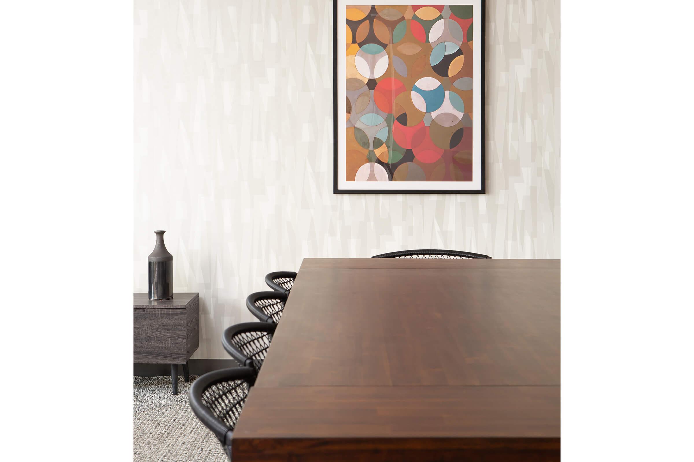 A modern conference room featuring a large wooden table with black woven chairs. A side table holds a dark vase. The wall is adorned with a colorful abstract art piece in a black frame, complementing the overall contemporary decor and light-colored textured wallpaper.