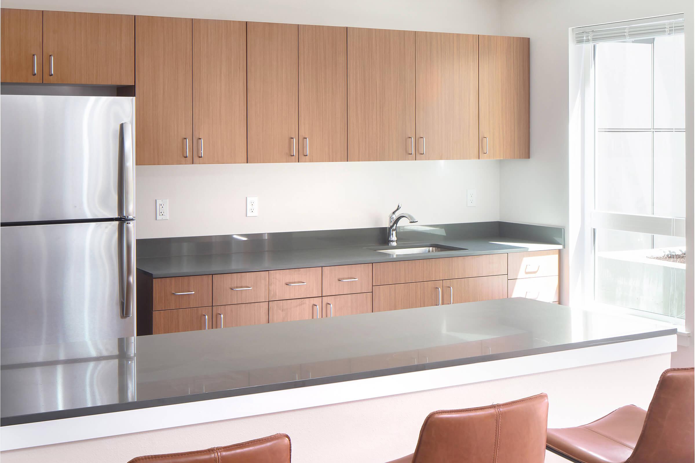 Modern kitchen featuring wooden cabinetry, a stainless steel refrigerator, and a sleek black countertop. The space includes a sink and minimalistic design with natural light coming in from a large window. Brown leather bar stools are positioned at the counter for seating.