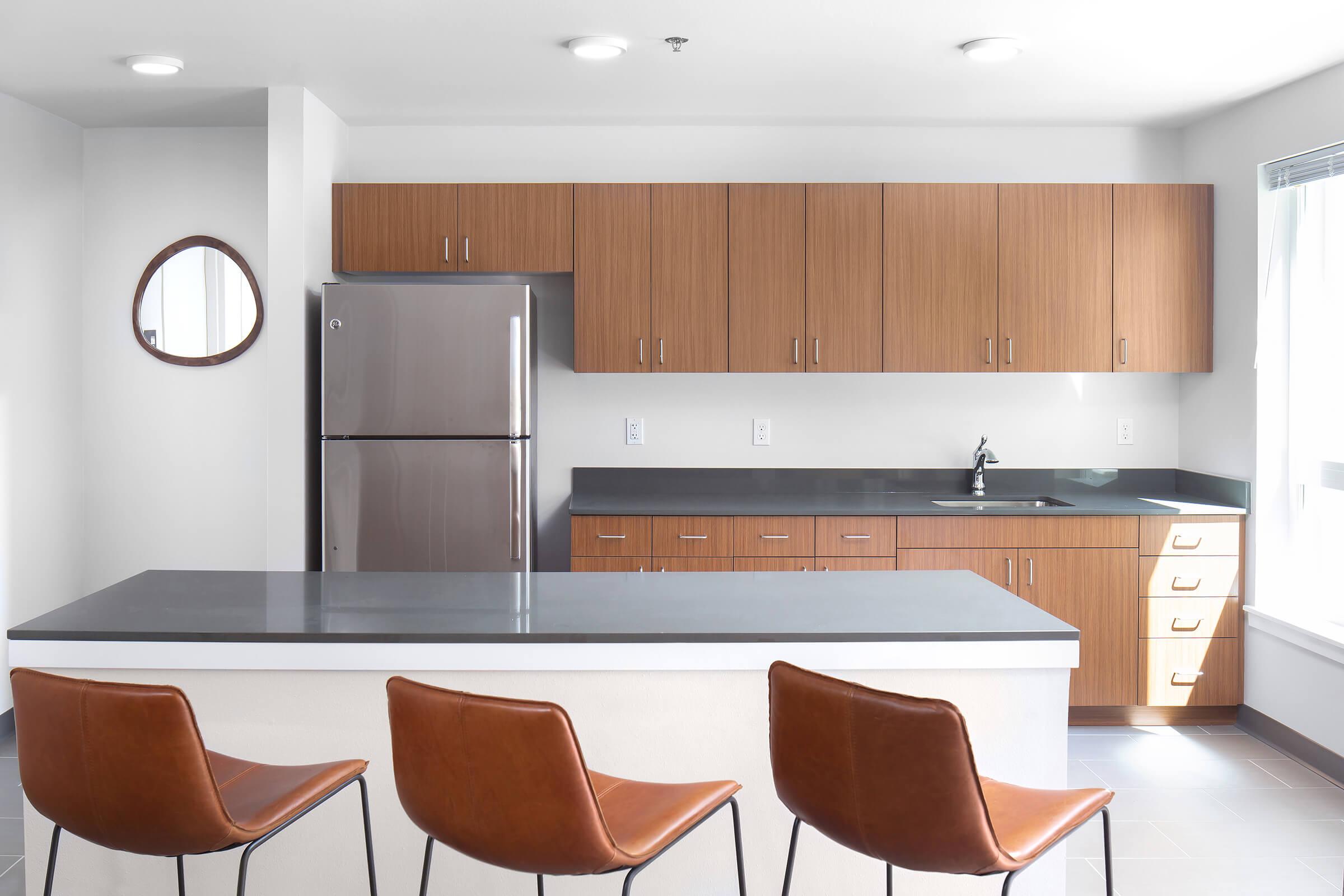 A modern kitchen featuring wood cabinets, stainless steel appliances, and a gray countertop. There are three brown leather bar stools at the kitchen island, and a round mirror is mounted on the wall. The space is well-lit with natural light coming through the window, creating a clean and contemporary atmosphere.