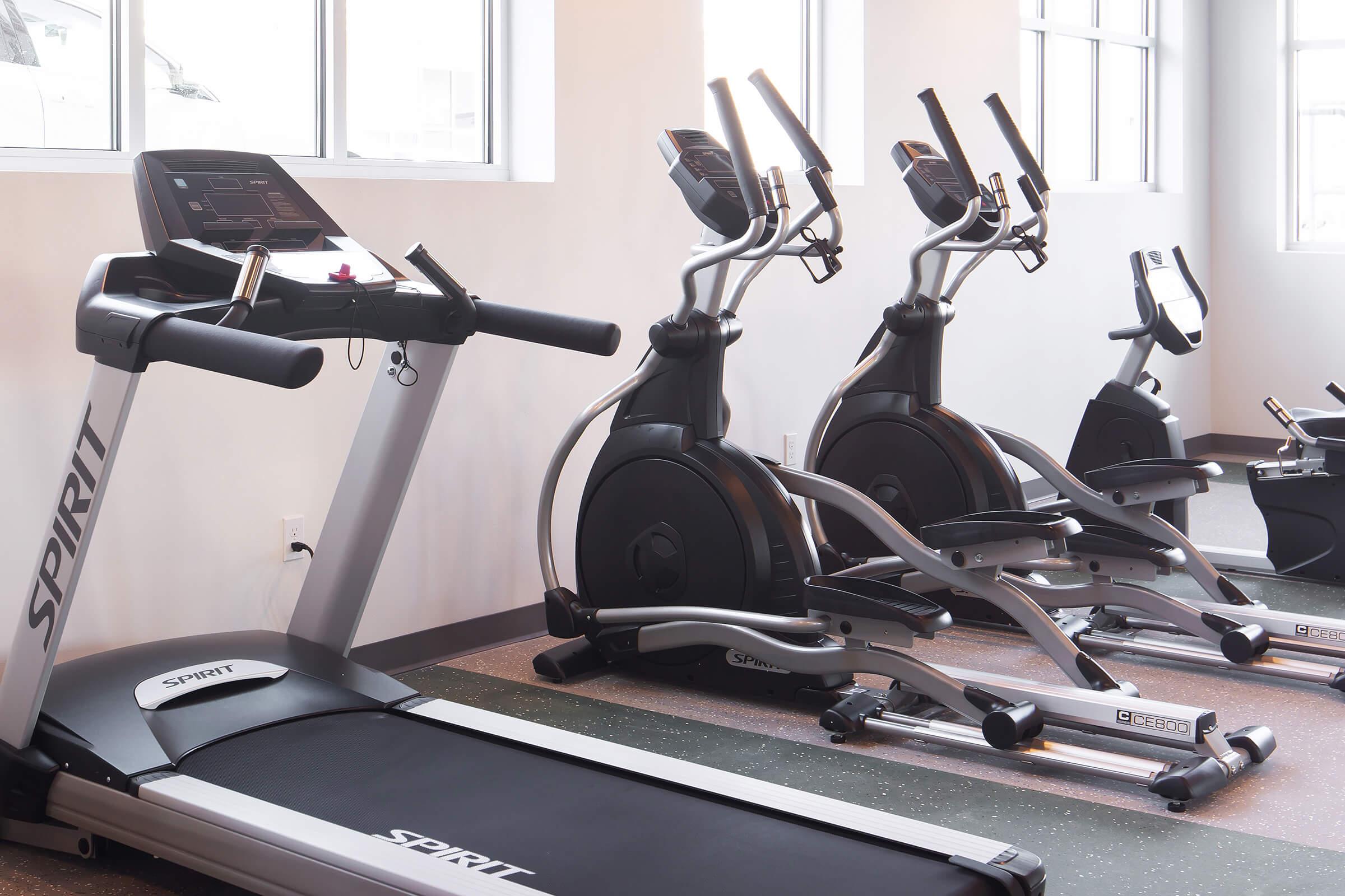 A modern gym equipment setup featuring a Spirit treadmill in the foreground and two elliptical machines in the background. The space is well-lit with large windows, creating an inviting atmosphere for exercise. The flooring appears multi-colored, adding a vibrant touch to the gym environment.