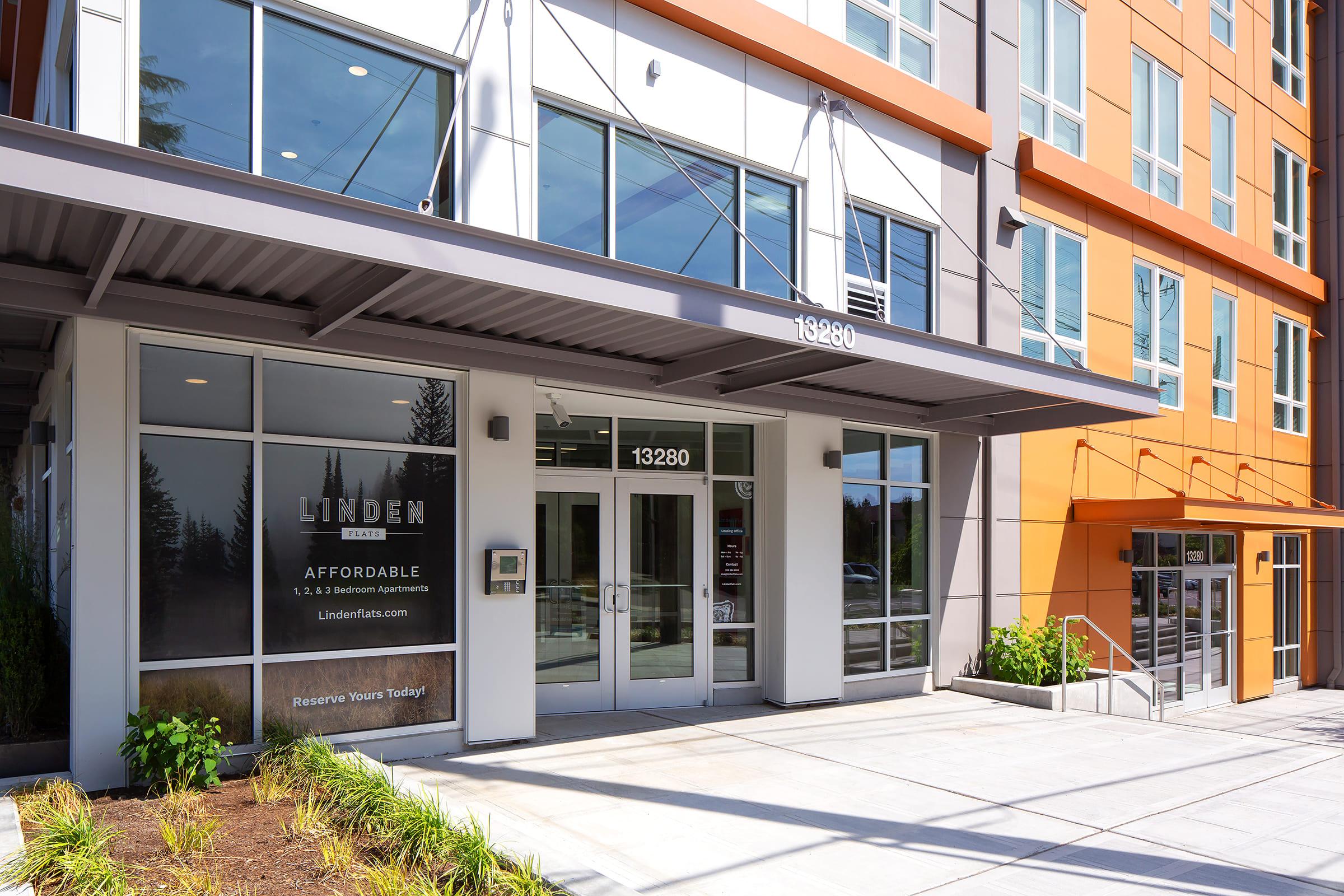 Exterior view of a modern apartment building named "Linden." The entrance features large glass doors and a covered awning. Adjacent signage promotes affordable housing with the text "Reserve Yours Today!" The building has a contemporary design with orange and gray accents.