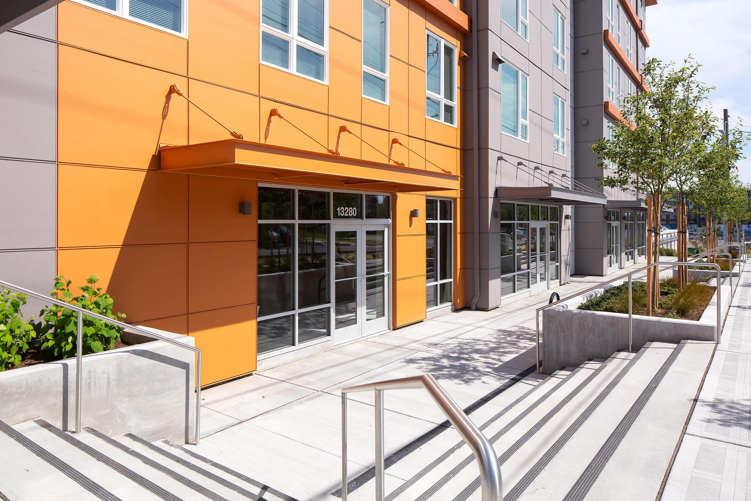 Modern building exterior featuring a vibrant orange and gray façade, with large glass windows and entrance. The area includes concrete walkways, steps with handrails, and landscaping with small trees and shrubs. Sunlight illuminates the scene, emphasizing the contemporary design.