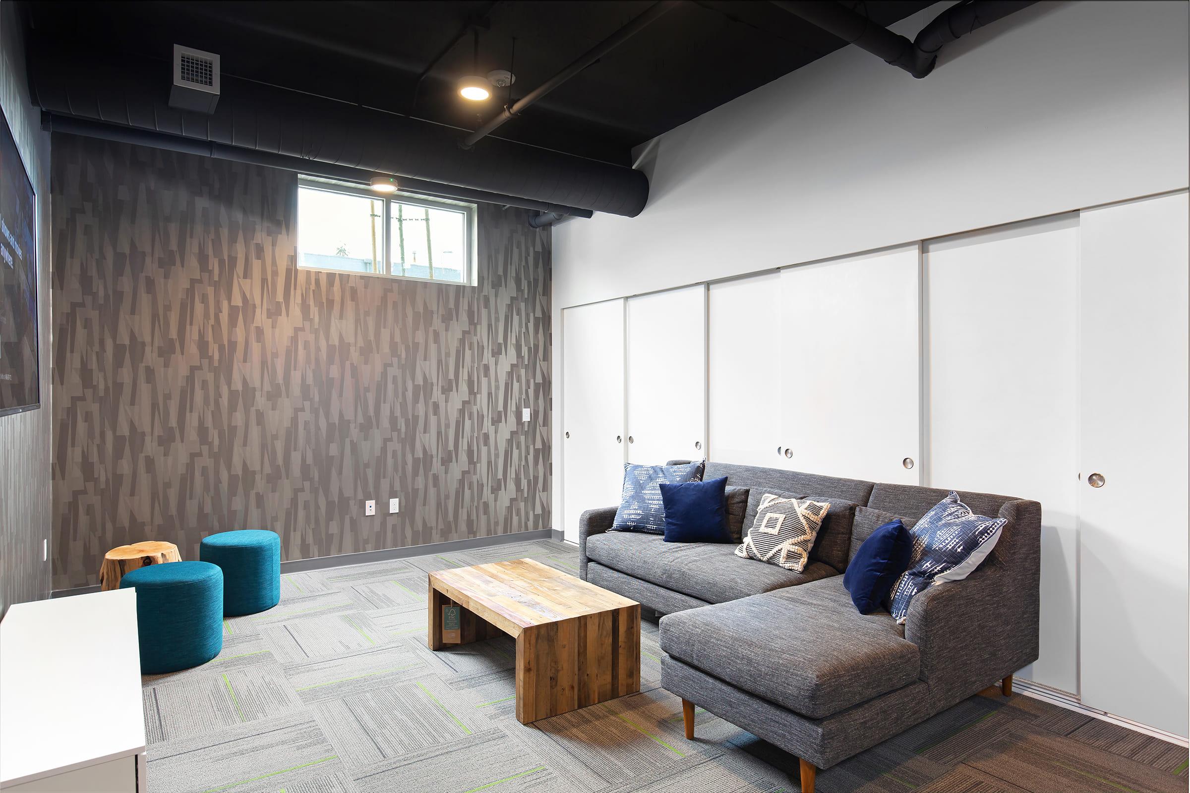 A modern lounge area featuring a gray sofa with decorative pillows, a wooden coffee table, two blue poufs, and a wall with a sleek pattern. The room has a large window and light-colored walls, creating a bright and inviting atmosphere.