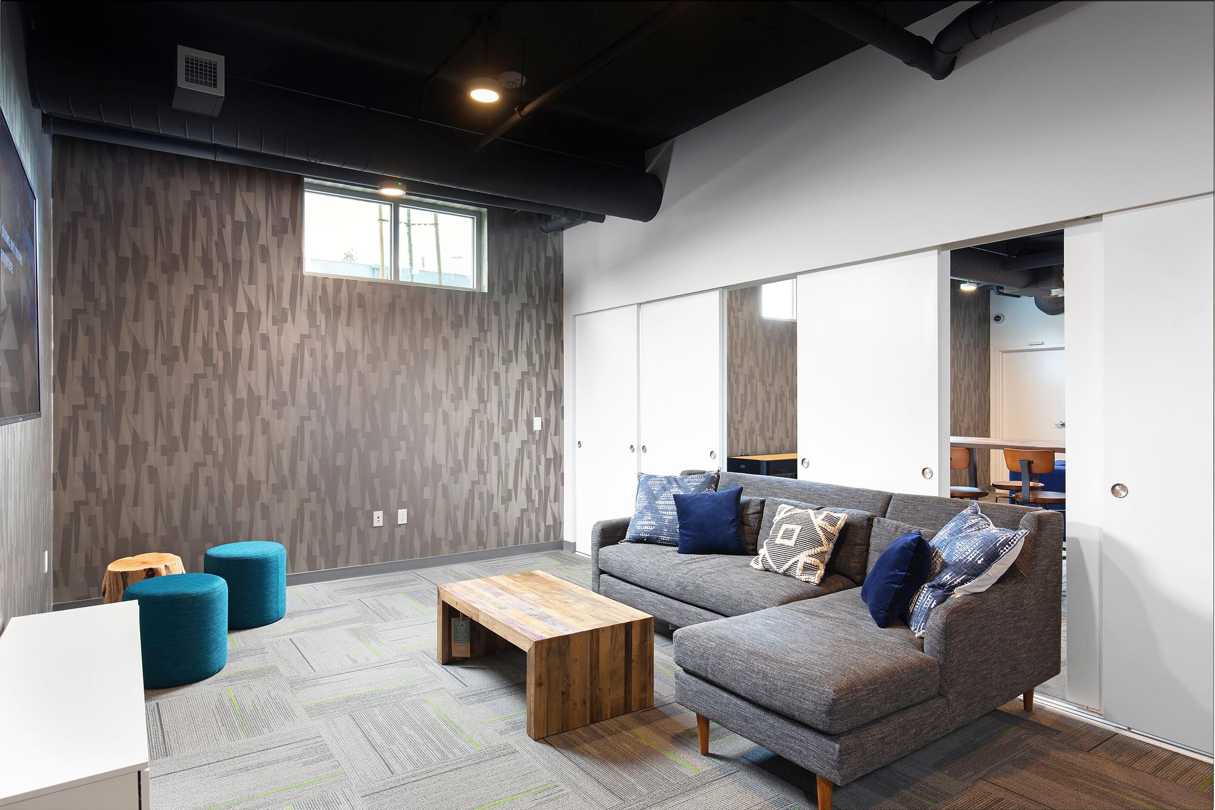 A modern lounge area featuring a gray sofa with decorative pillows, a wooden coffee table, and blue stools. The walls have a textured pattern, and there is a window allowing natural light. In the background, there are white-panel sliding doors and a glimpse of another room.