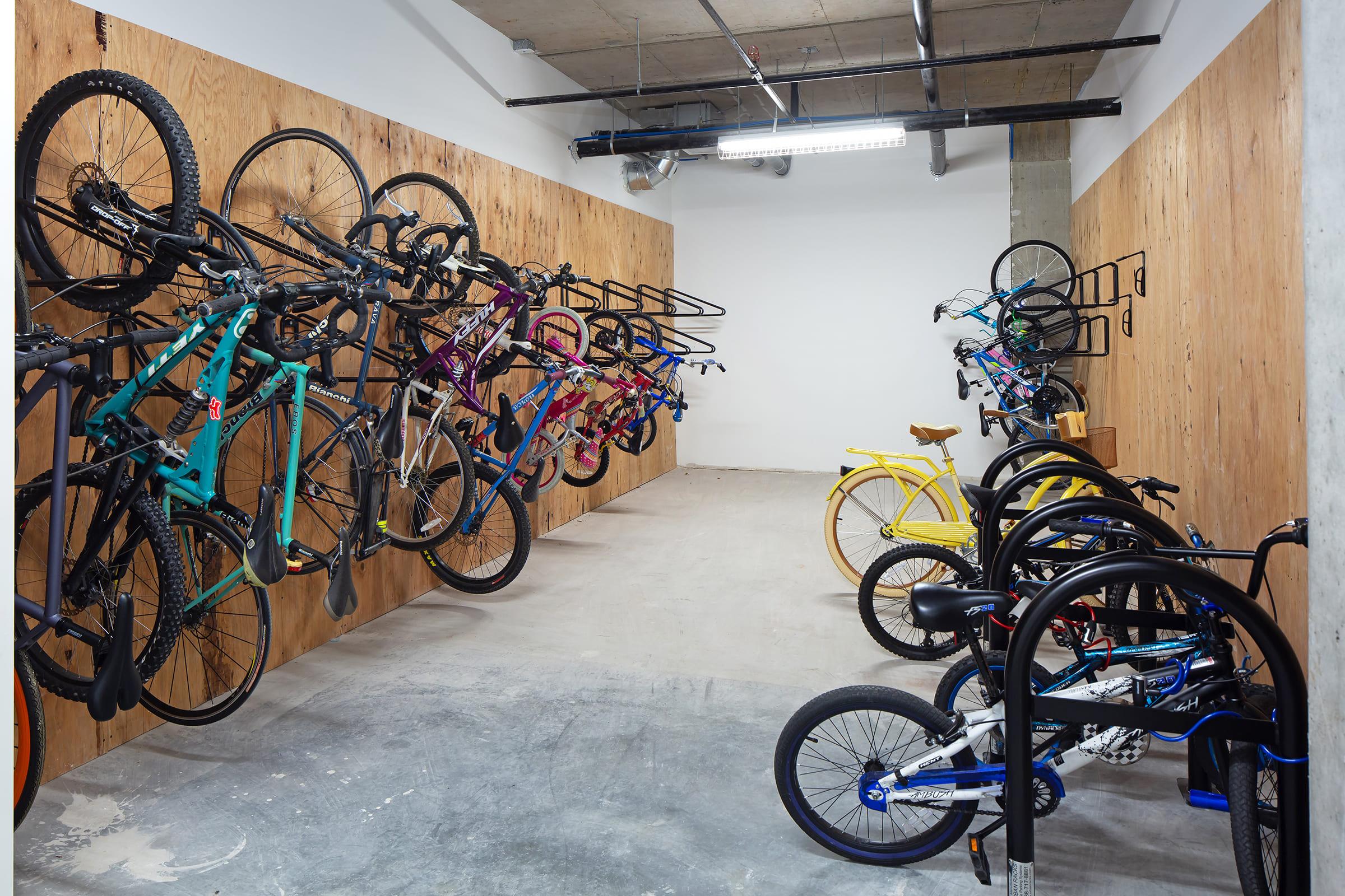 A modern bicycle storage area featuring various bicycles hanging on wooden walls and several bikes parked on the floor. The bikes come in different colors and styles, including mountain bikes and cruisers, with a clean, minimalistic design and concrete flooring.