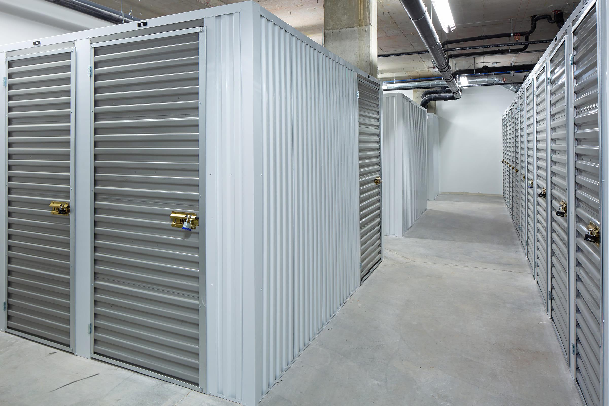 A row of metal storage units with metal doors and locks, arranged in a well-lit indoor facility. The corridor between the units features a concrete floor and exposed pipes on the ceiling, creating a minimalist and functional storage environment.