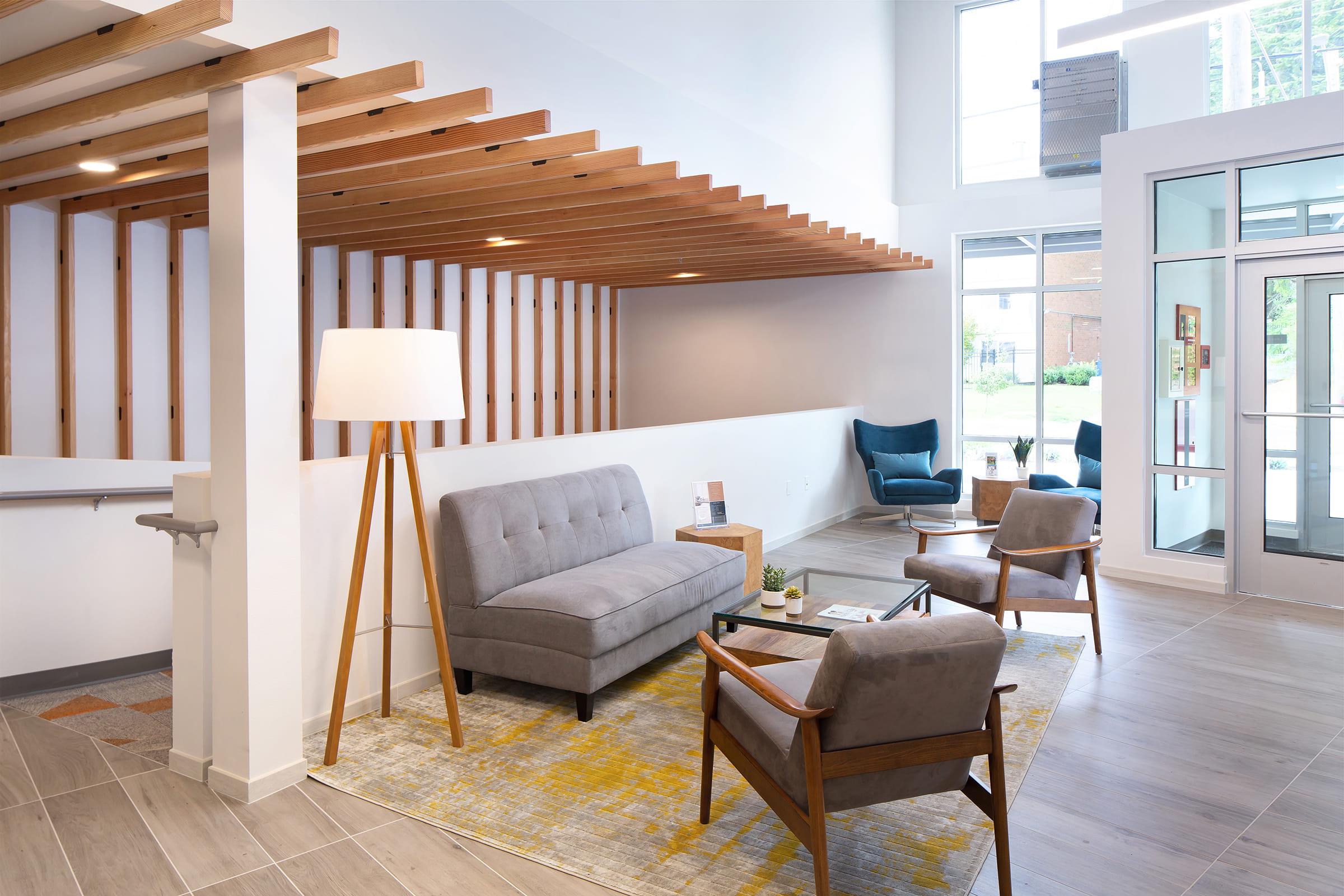 A modern lobby area featuring a gray sofa, two wooden armchairs, a floor lamp, and a table with decor. The space is illuminated by natural light from large windows, with a stylish wooden ceiling and neutral color palette, creating a welcoming atmosphere.