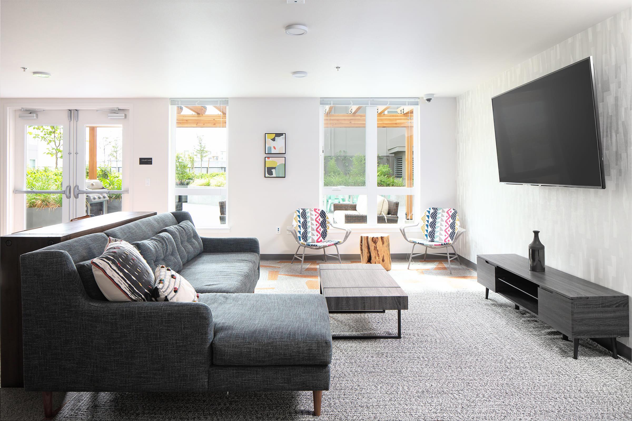 A bright and modern living room featuring a gray sectional sofa with decorative pillows, a wooden coffee table, and a sleek TV stand. Large windows let in natural light, and two stylish chairs sit beside a glass door leading to an outdoor area. The walls have a textured design, adding a contemporary touch.