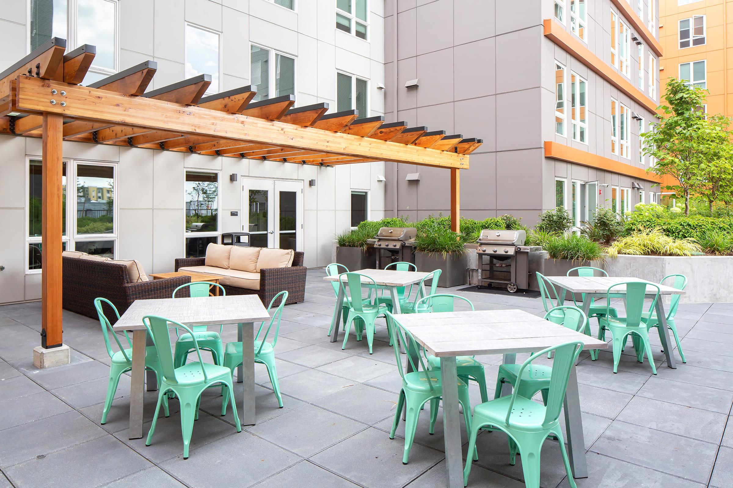 Outdoor patio area featuring modern furniture. There are several mint green tables and chairs, a shaded seating area with a sofa under a wooden pergola, and two barbecue grills. The space is surrounded by lush greenery and contemporary apartment buildings.