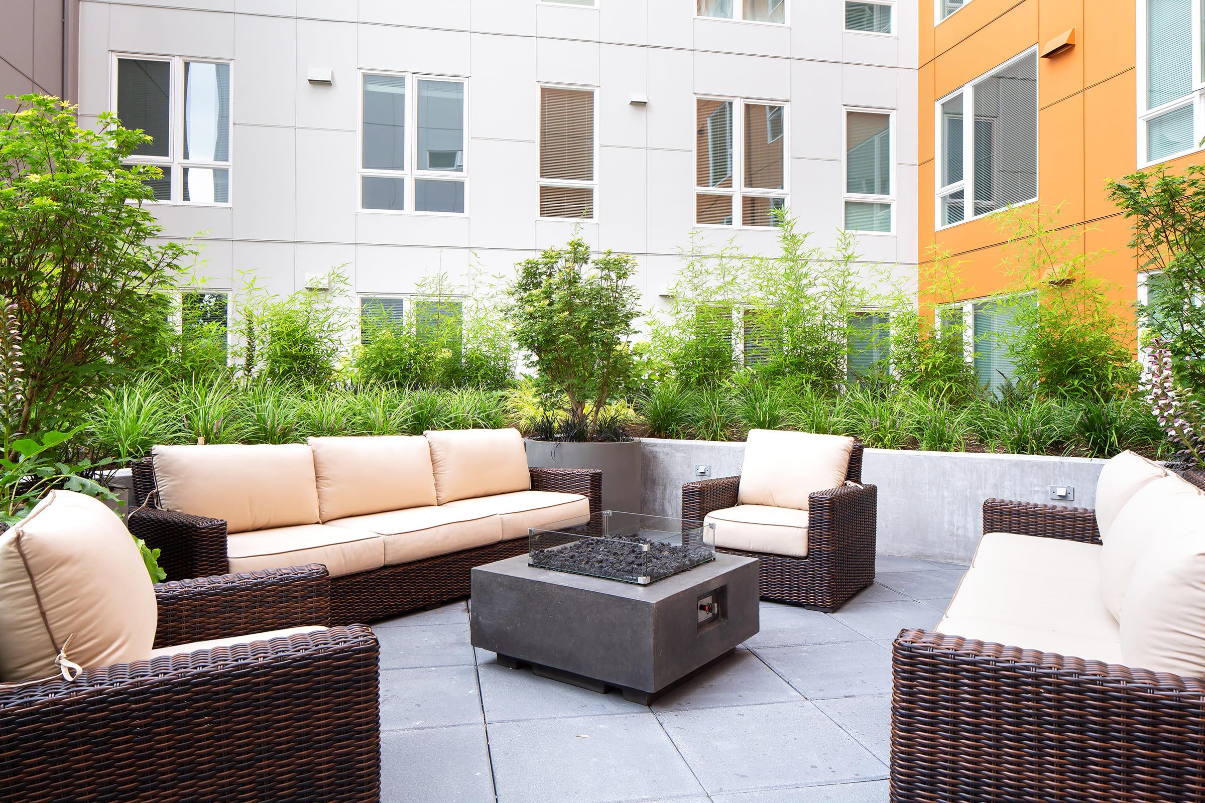 A modern outdoor lounge area featuring comfortable beige sofas and armchairs made of wicker. A square fire pit sits in the center on a paved surface, surrounded by lush greenery and low plants, with buildings visible in the background. The setting offers a cozy atmosphere for relaxation.