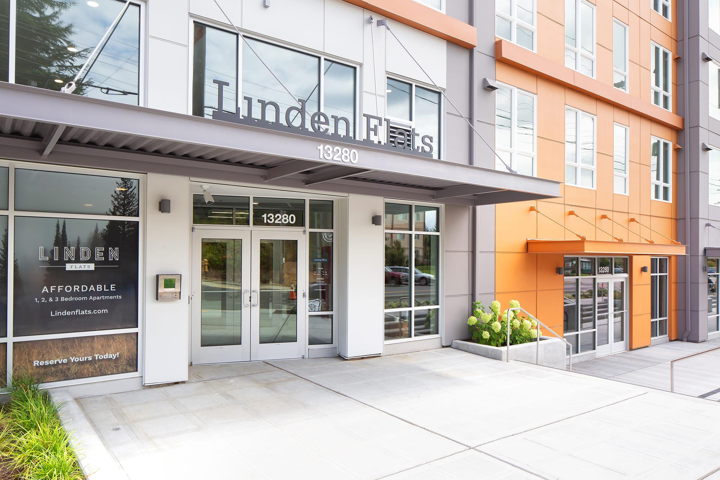 Modern apartment building exterior featuring large glass entrance doors, the name "Linden Flats" prominently displayed above, and a spacious entrance pathway. The building has a mix of colors, including beige and orange, and is surrounded by greenery. A sign promotes reserving apartments.