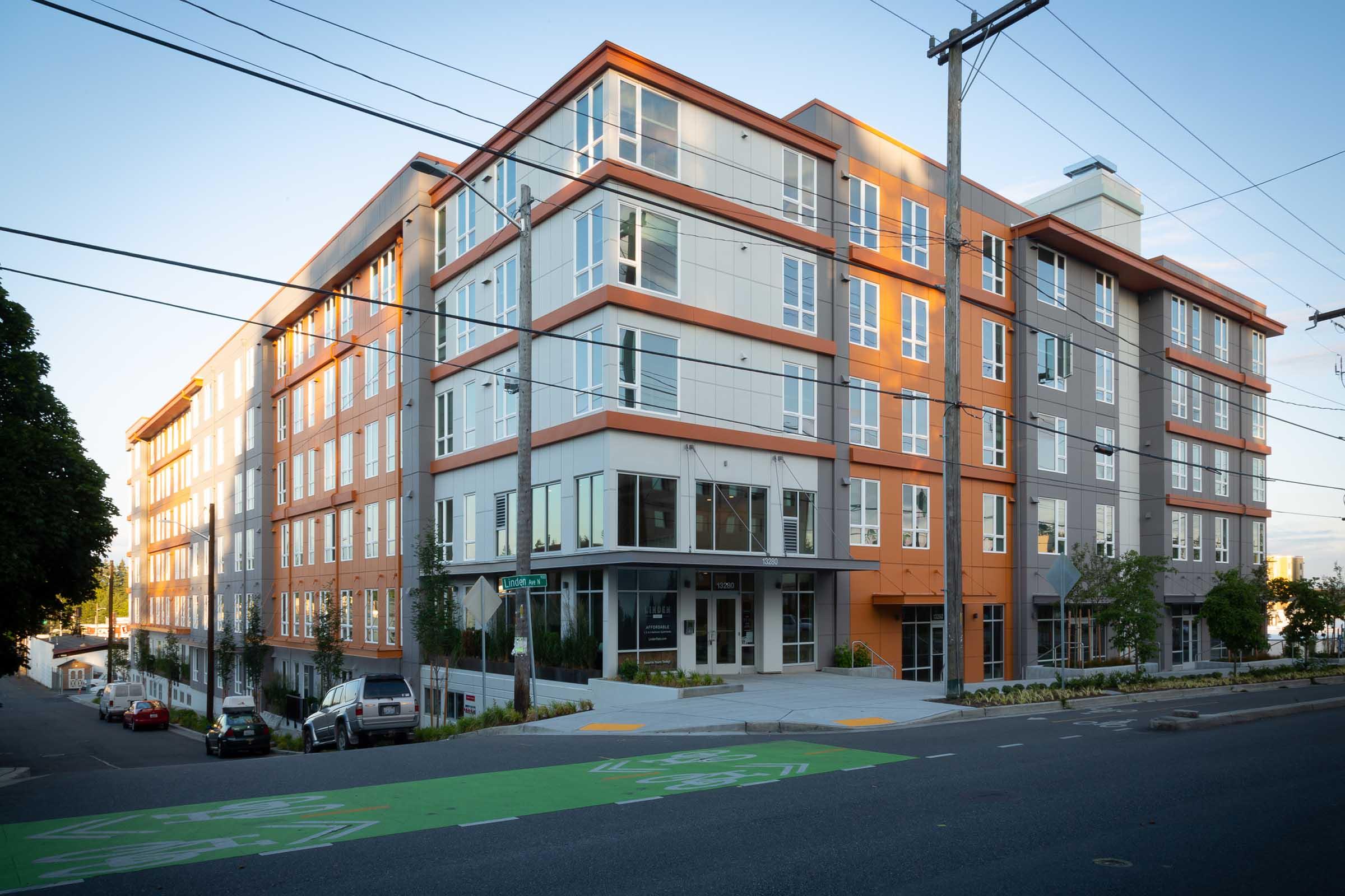 A modern multi-story residential building with a mix of orange and gray facade. Large windows and a spacious entrance are visible. The street in front features green bike lanes and parked cars. Power lines are overhead, and the setting appears to be in an urban neighborhood.