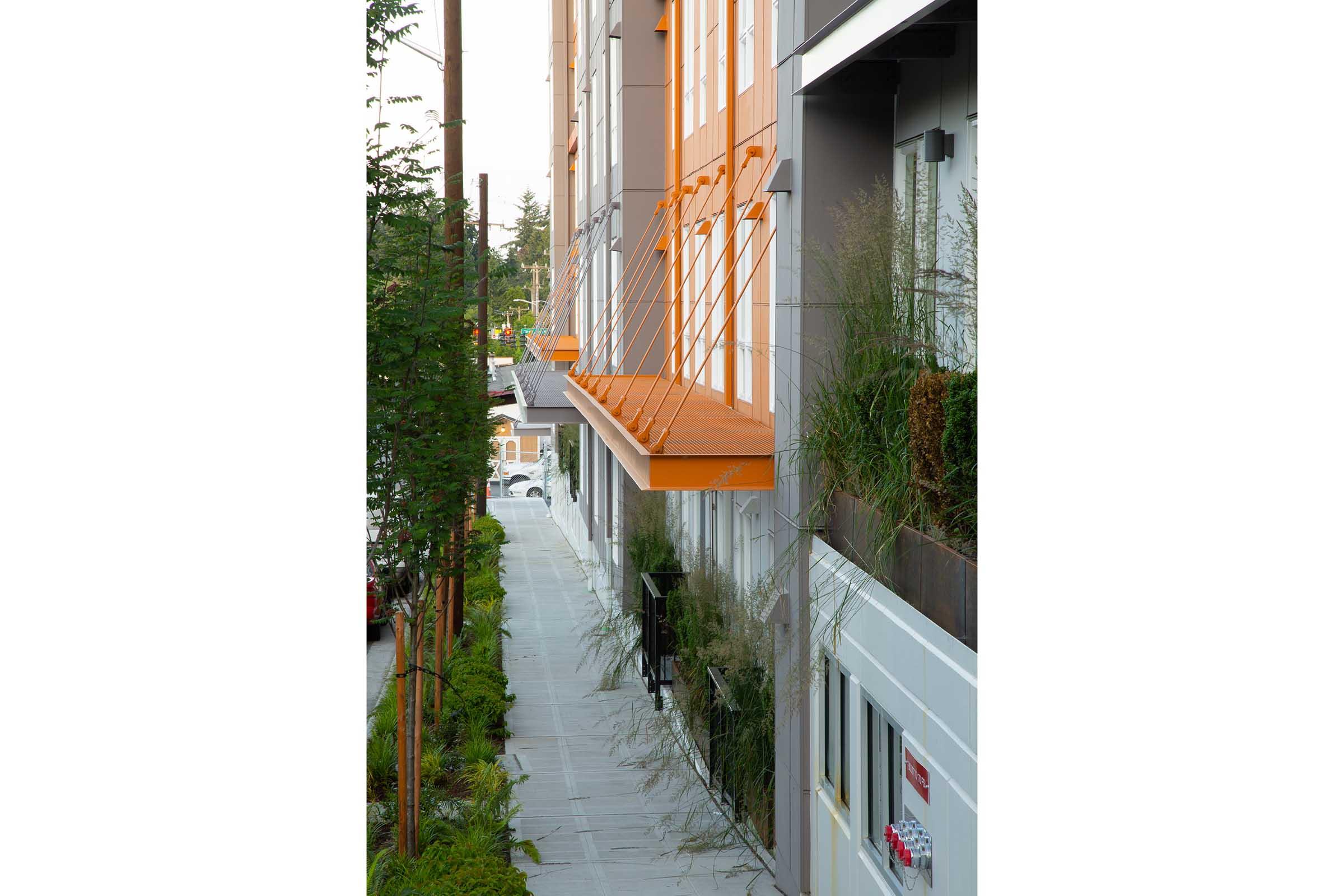 A modern building features orange balconies extending over a sidewalk lined with greenery. The surrounding area includes small trees and plants, creating a visually appealing urban environment. The scene captures a clean and contemporary architectural design in a pedestrian-friendly setting.