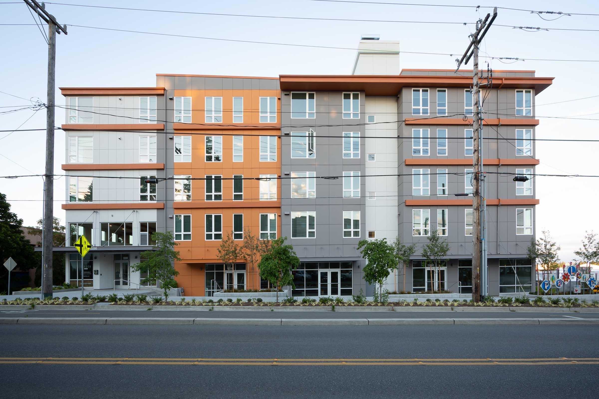 Modern multi-story building featuring a mix of orange and gray facades. Large windows adorn each floor, and trees are planted in front. Power lines run overhead, and a crosswalk sign is visible to the left. The scene is set along a road, showcasing urban architecture.