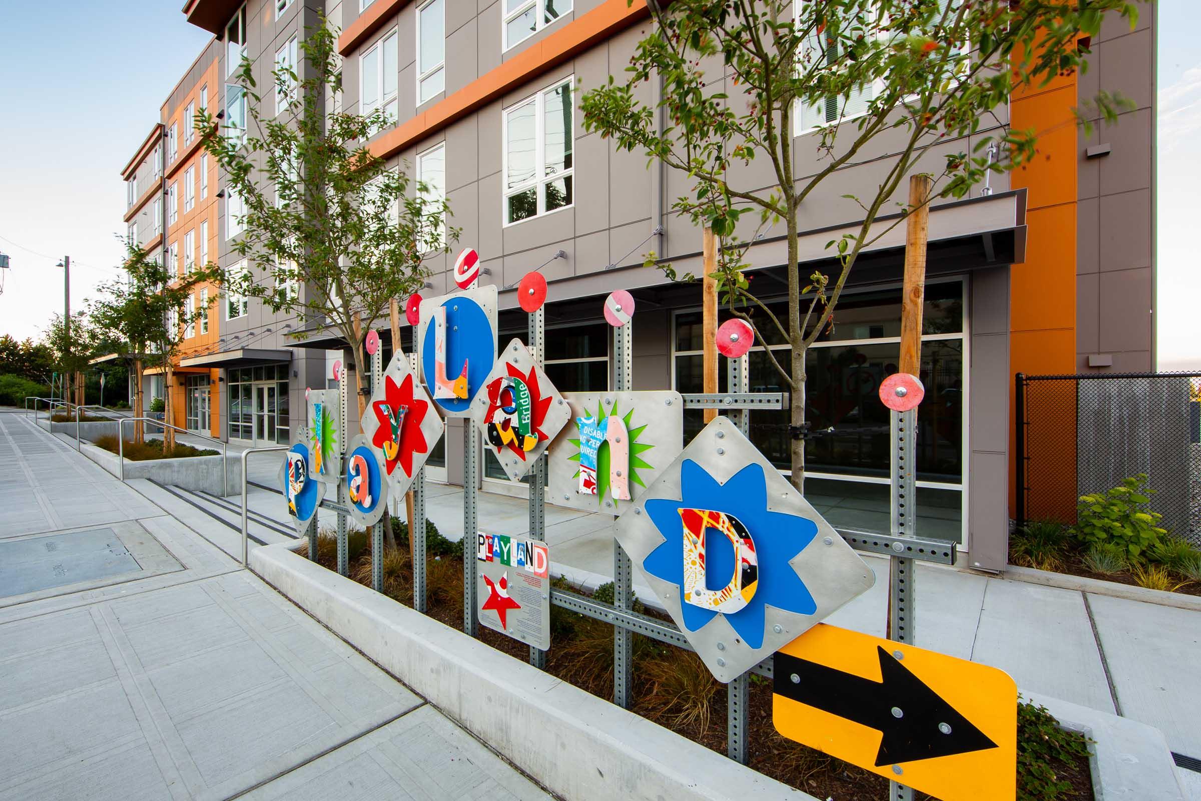 Colorful outdoor display featuring various playful signs and letters arranged on a fence, surrounded by small trees, with a modern apartment building in the background. The signs include shapes like stars and arrows, creating a vibrant and inviting atmosphere. Sidewalk path visible in the foreground.