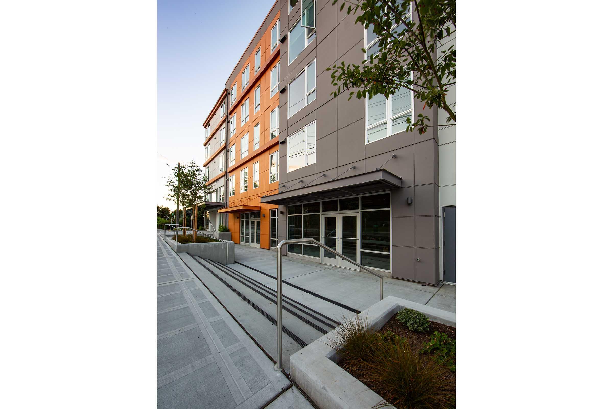 A modern multi-story building with a mix of orange and gray exterior panels. The entrance features large glass doors and a covered area with sleek architectural lines. Surrounding the entrance are landscaped areas with plants and a smooth walkway leading to the building. The scene is set in the early evening light.