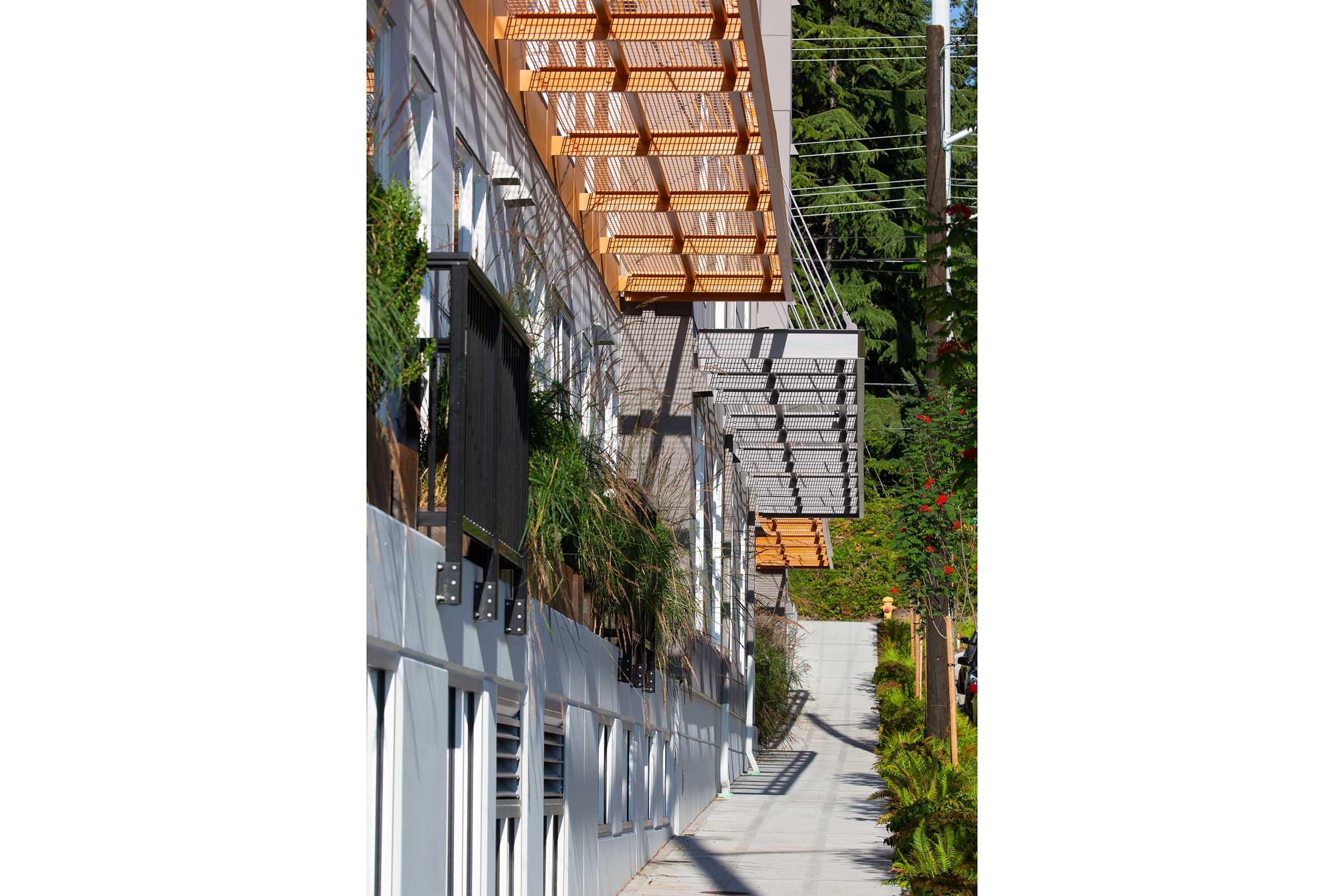 A walkway alongside a modern building featuring large windows and metal awnings. The exterior is adorned with greenery and plants, and a pathway runs alongside the structure, leading towards a tree-lined area. The image captures a sunny day, highlighting architectural details and landscaping.